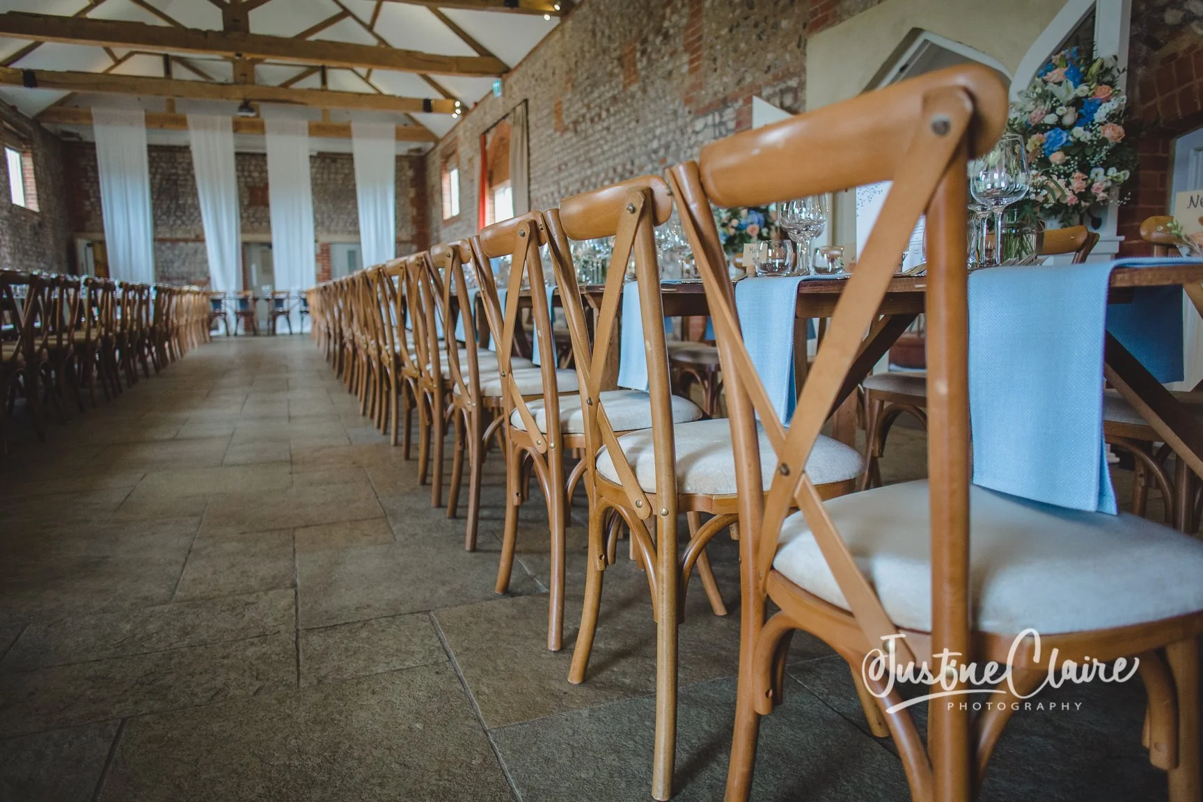 Rows of wooden chairs aligned along a long banquet table in a rustic farbridgewith brick walls and high wooden ceilings, decorated with flowers and table settings.