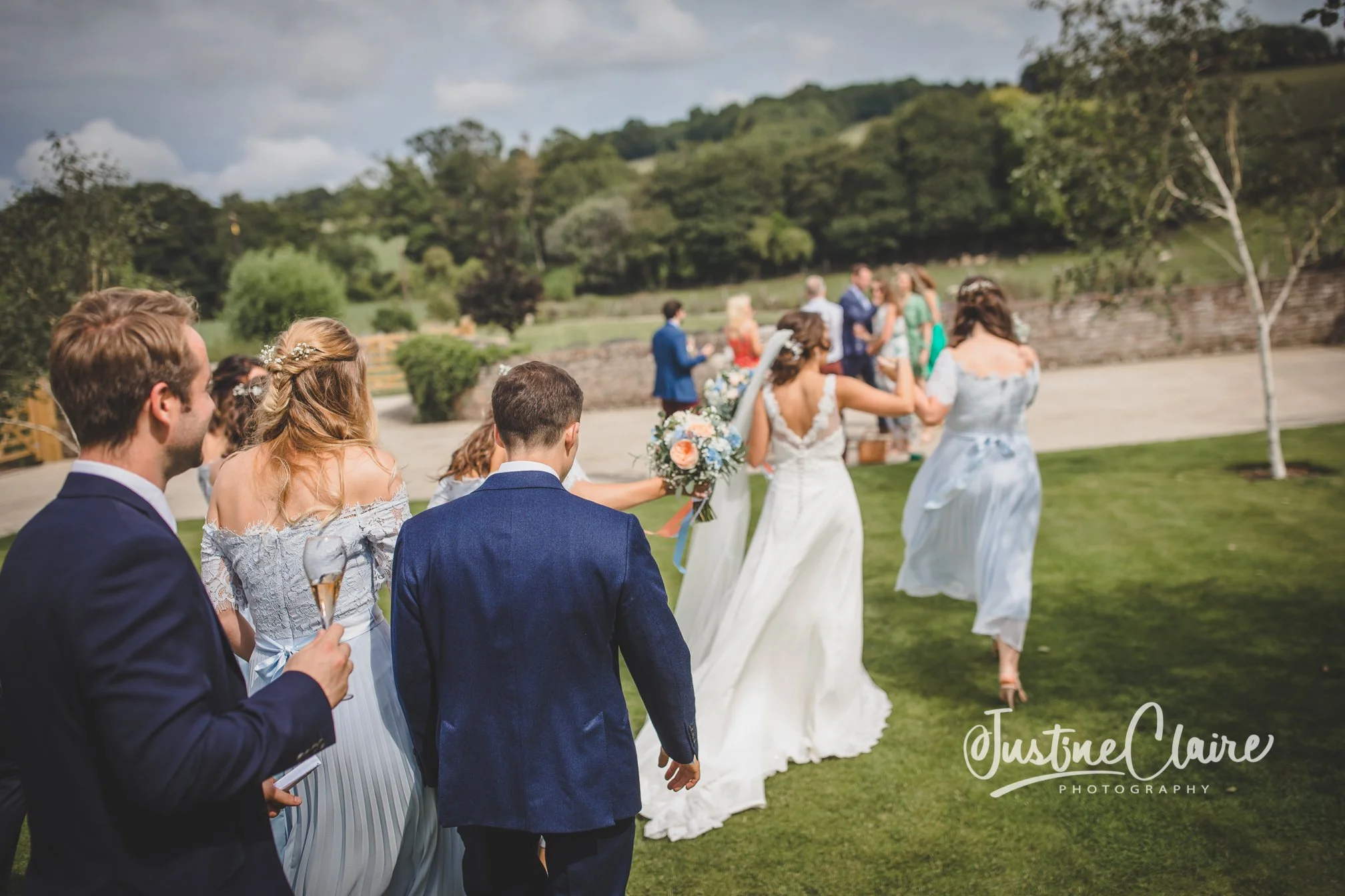 Wedding ceremony outdoors with guests walking on a grassy area, with a bride holding a bouquet leading the group, surrounded by friends and family, in a scenic landscape with trees and hills in the background.