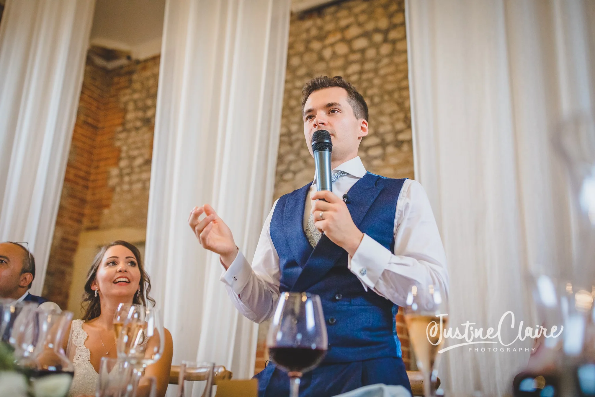 A man in a blue vest and white shirt giving a speech at a wedding reception, holding a microphone. A woman in a wedding dress looks up at him, smiling, sitting at a table with wine glasses. The background features tall white curtains and a brick wall