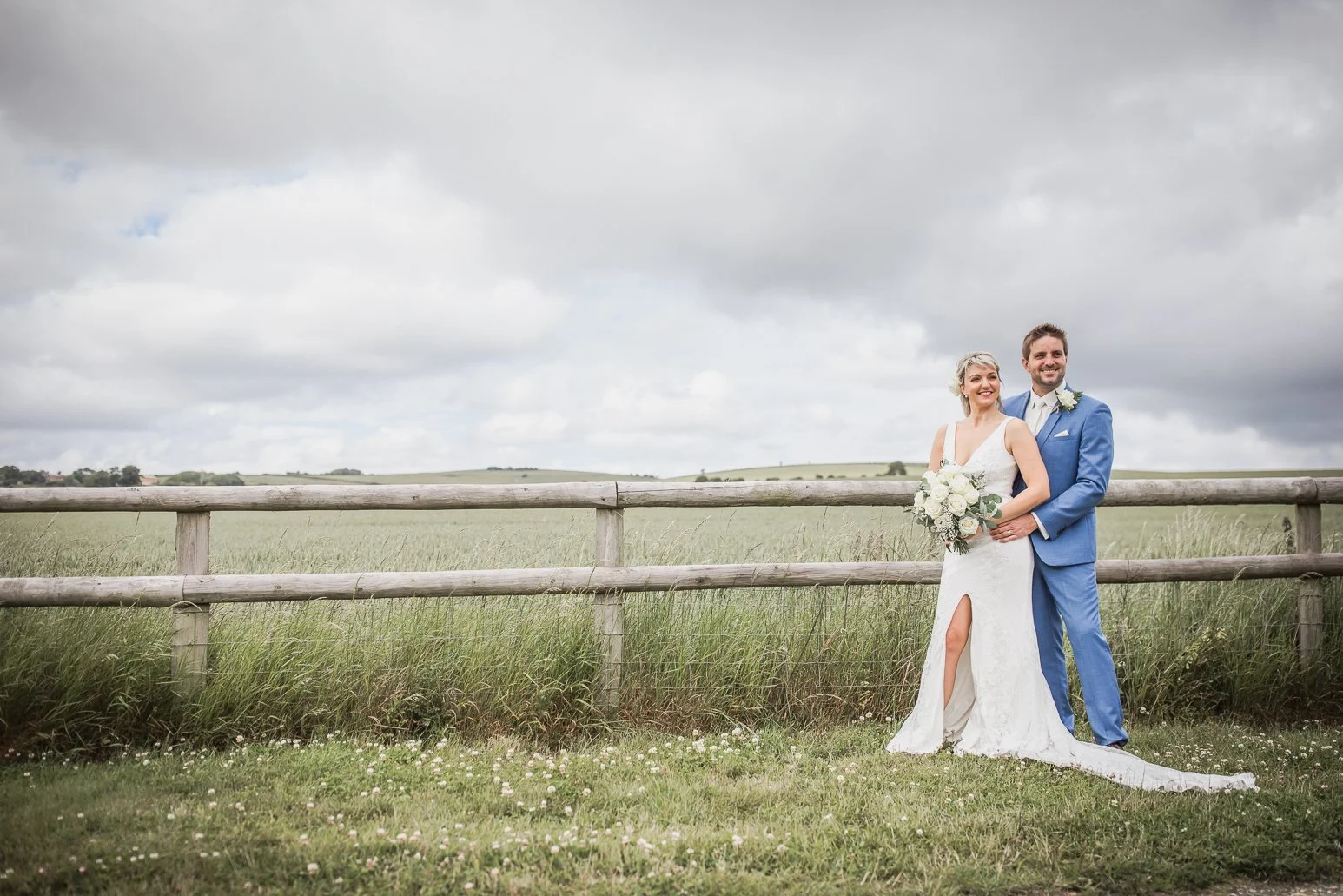 Bride and groom standing outdoors in front of a wooden fence, smiling and holding a bouquet of white flowers, with cloudy sky and green fields in the background.