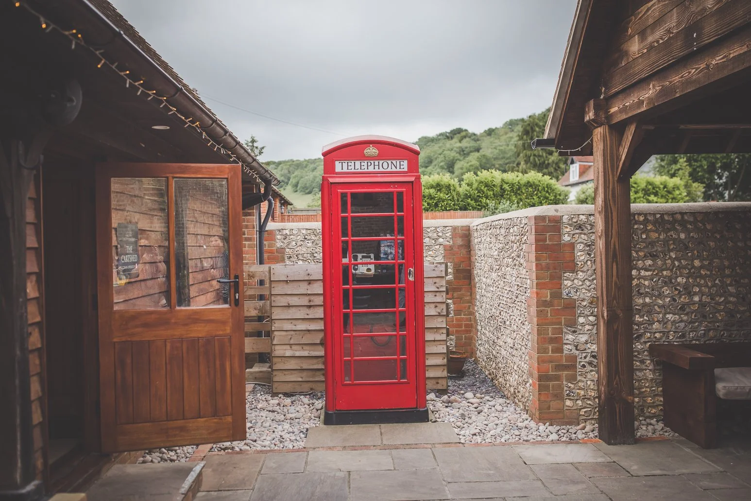 A classic red British telephone booth centered between two wooden structures with tiled roofs, in an outdoor area with stone paving, a fence, and a background of green hills under an overcast sky.