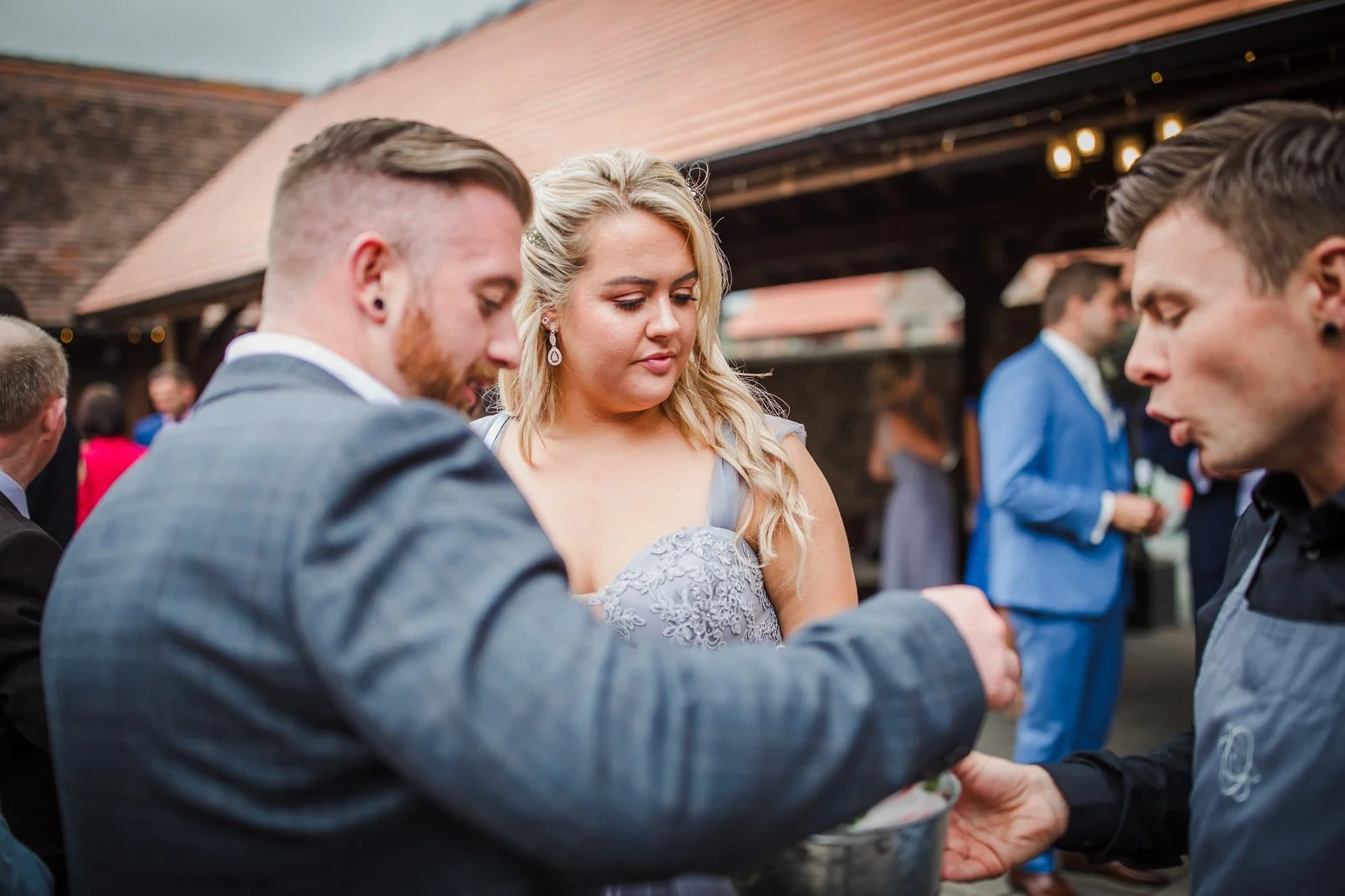 A group of people dressed in formal attire at an outdoor wedding, with a woman in a gray lace dress and two men in suits, engaged in a ceremonial activity.