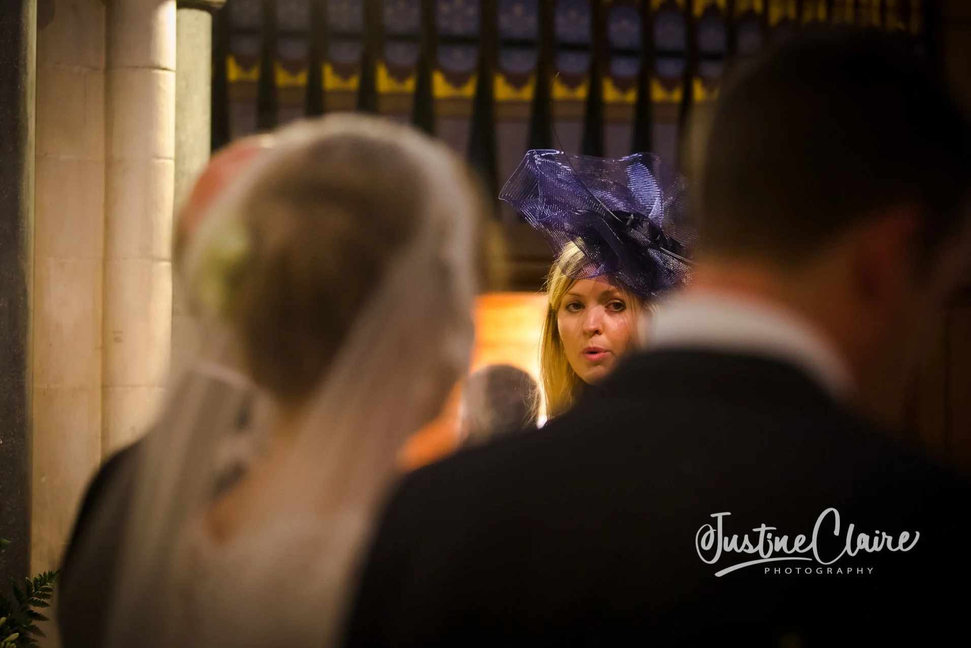 A woman wearing a large purple fascinator hat looks over her shoulder at two men in formal attire, with one of them wearing glasses, during a social event in a warmly lit room with wooden and stone elements.