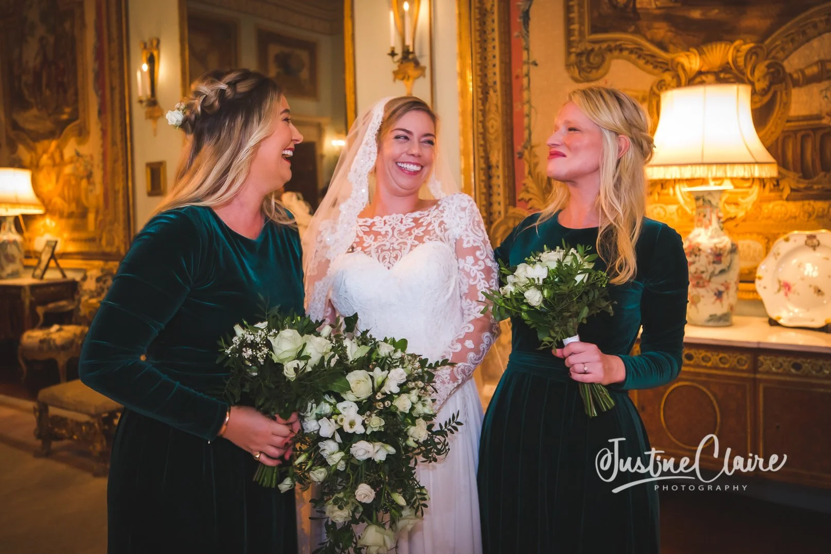 Three women at a wedding, with the bride in the middle wearing a lace wedding dress and veil, holding a bouquet of white flowers, smiling, and two bridesmaids on either side wearing dark green velvet dresses, also holding bouquets of white flowers, i