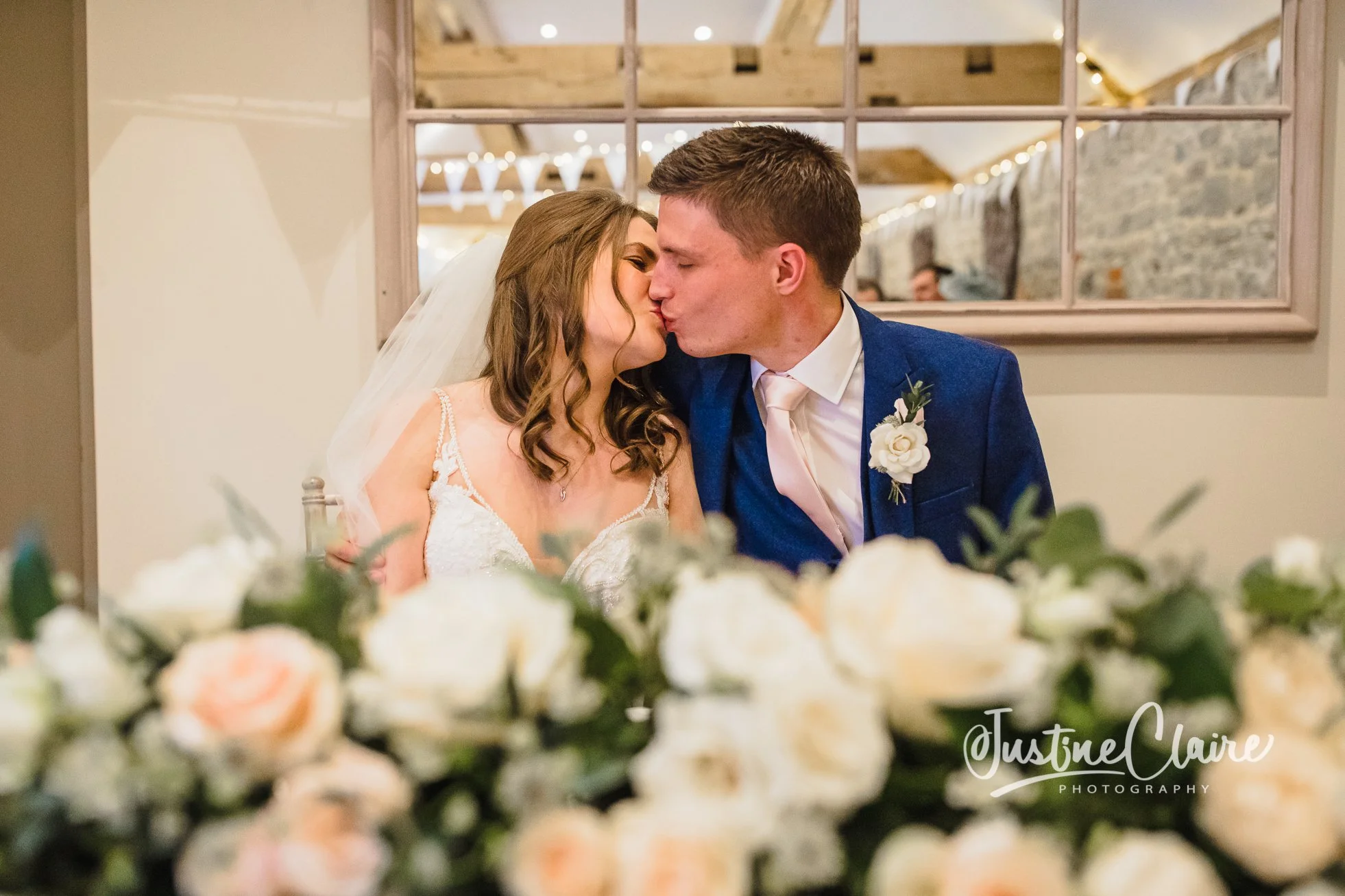 A bride and groom share a kiss at their wedding reception, sitting behind a table decorated with white and peach roses and greenery, with a mirror and string lights in the background. Fitzleroi barn wedding photographers