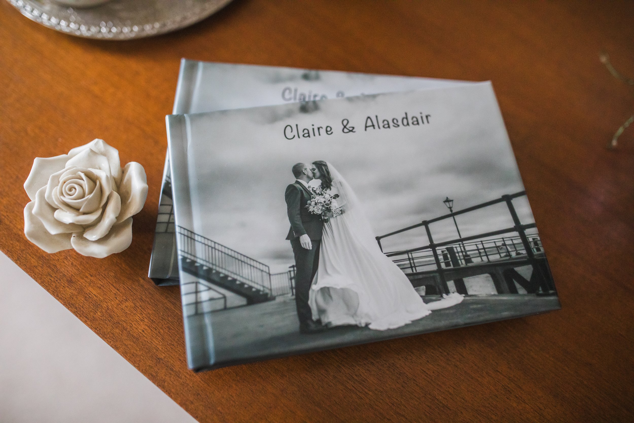 A wedding photo album with the names Claire & Alasdair on the cover, featuring a black and white photo of a bride and groom kissing on a pier, placed on a wooden surface alongside a white ceramic rose. 15 x 20 graphistudio parent book small