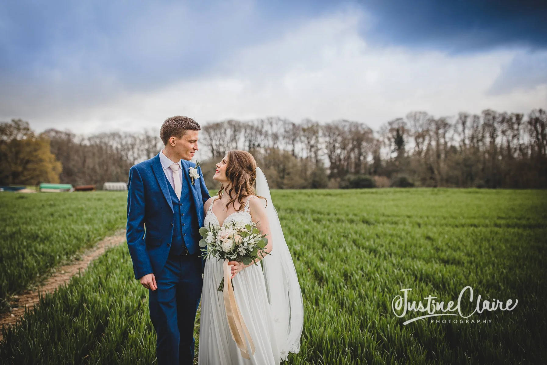 A bride and groom standing in a grassy field, gazing at each other, with trees and a cloudy sky in the background.