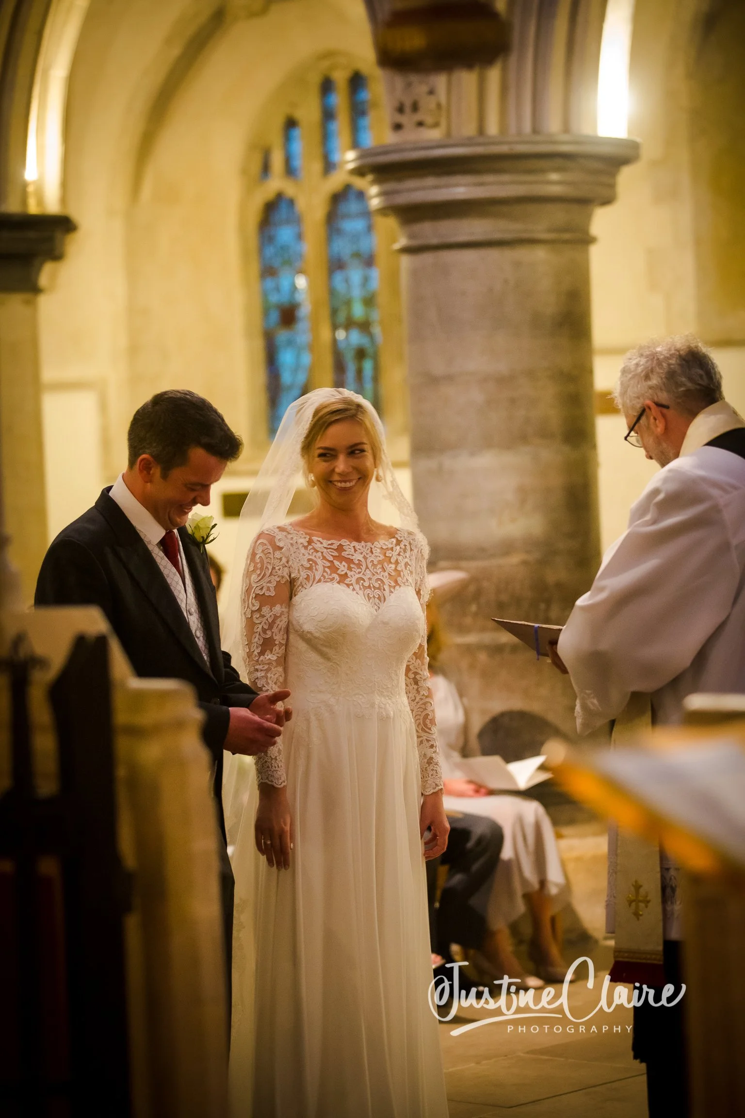 A bride and groom standing in a church during their wedding ceremony, with the officiant reading from a book. The bride is smiling, wearing a lace wedding dress with a veil, and the groom is dressed in a suit with a boutonnière. The church has arched