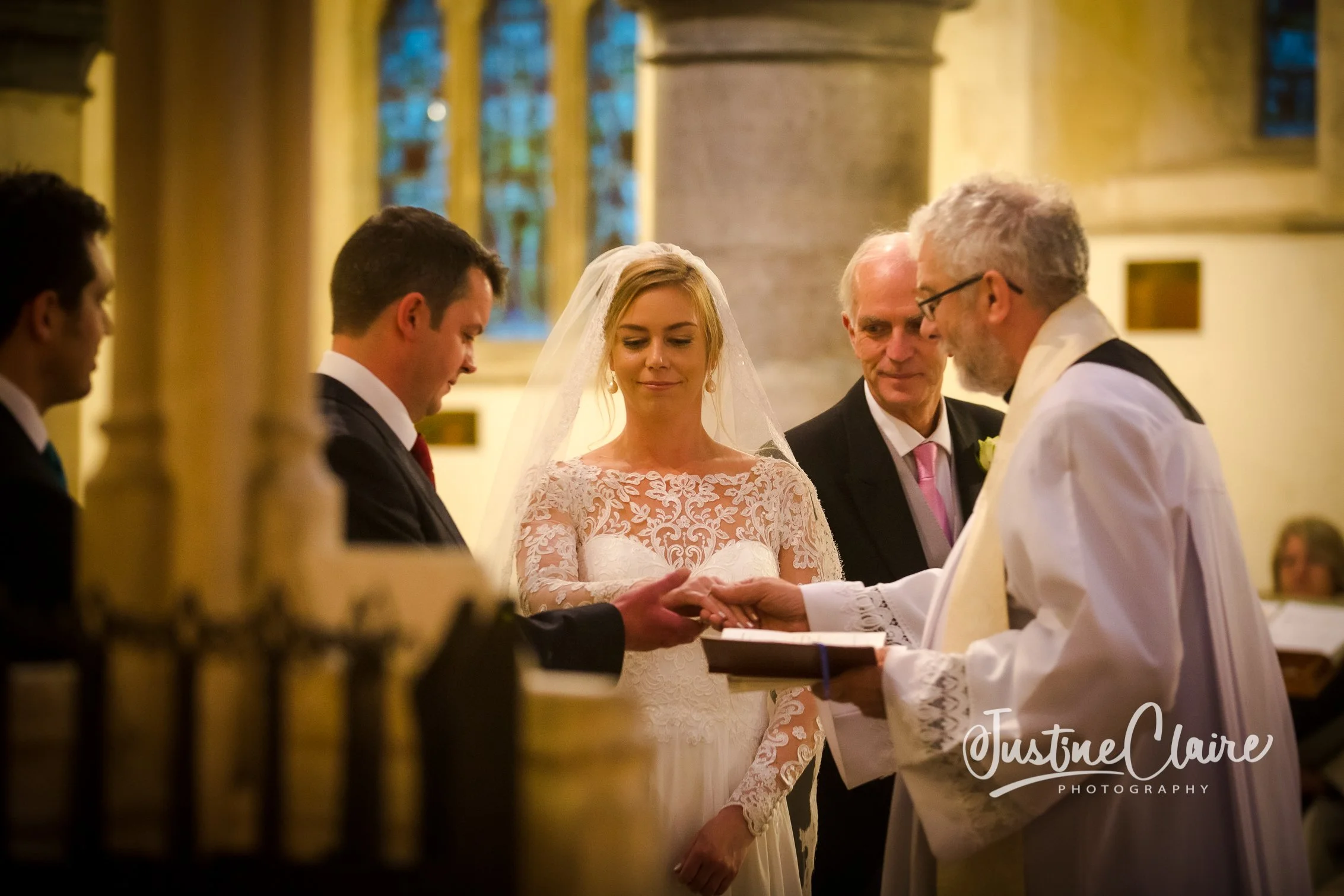 A wedding ceremony taking place inside a church, with a bride and groom holding hands in front of a priest, flanked by two men, possibly groomsmen, during an indoor ceremony with stained glass windows in the background.