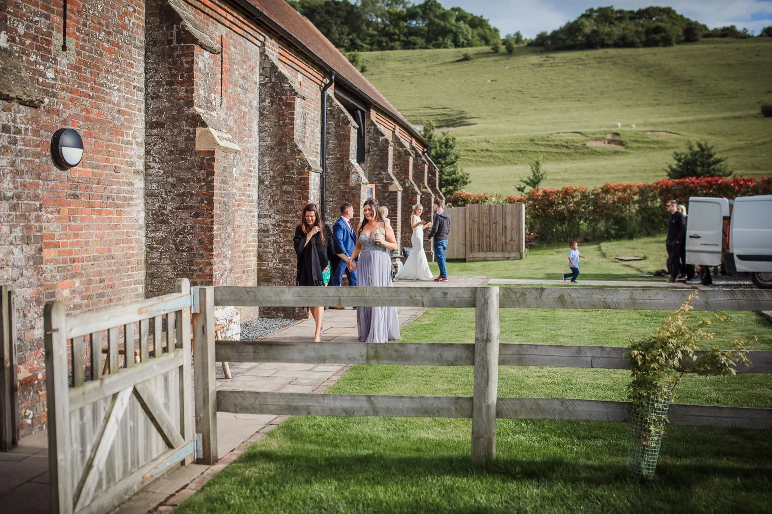 People gathered outside a brick building with a green hillside in the background, some dressed in formal attire, including a bride in a white wedding dress and a groom in a suit.