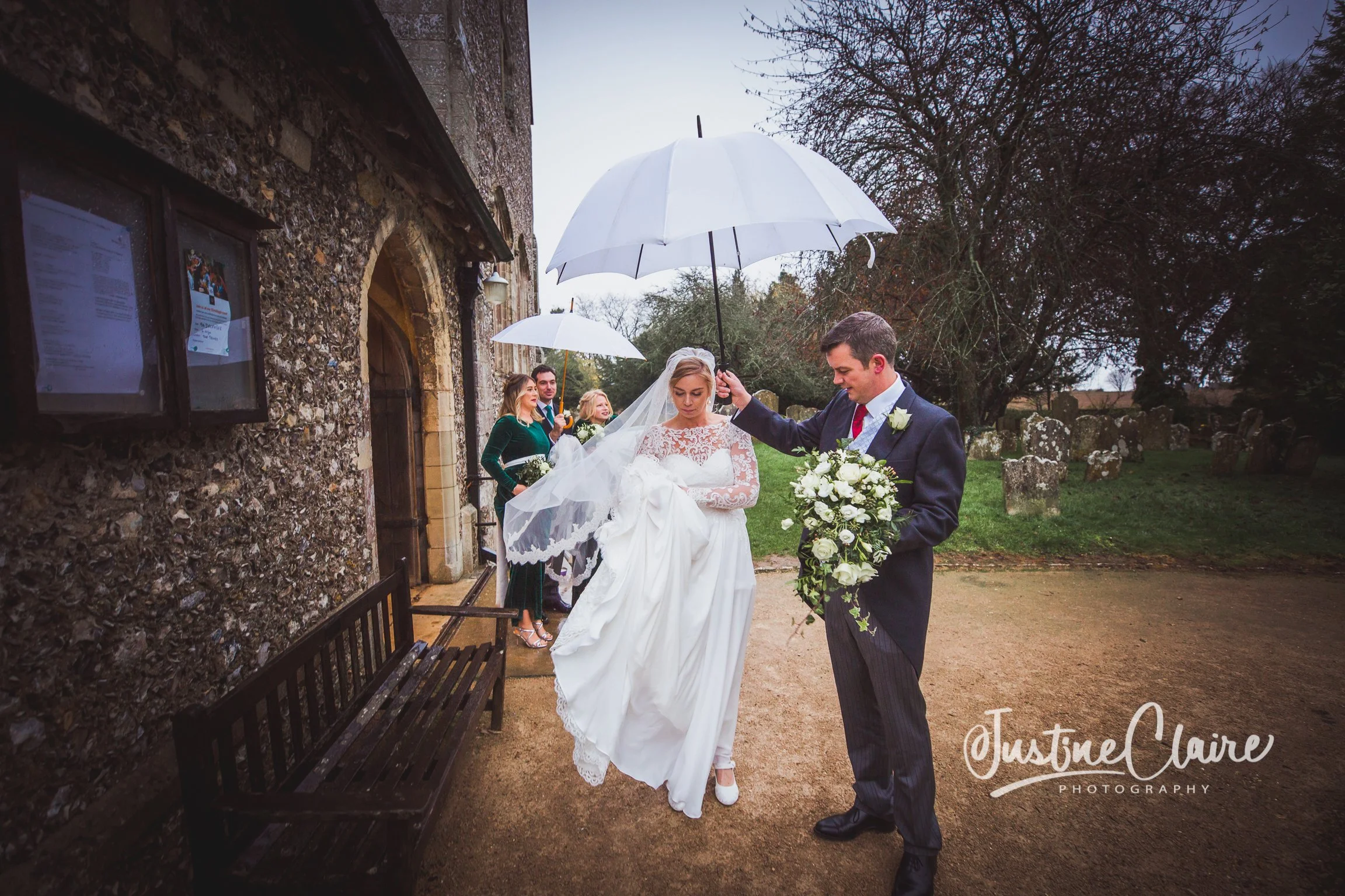 A wedding scene outside a church, with a bride in a white wedding dress and veil walking on a dirt path, and a groom in a navy suit holding a bouquet of white flowers. The groom is holding a white umbrella over the bride. Three women and one man stan