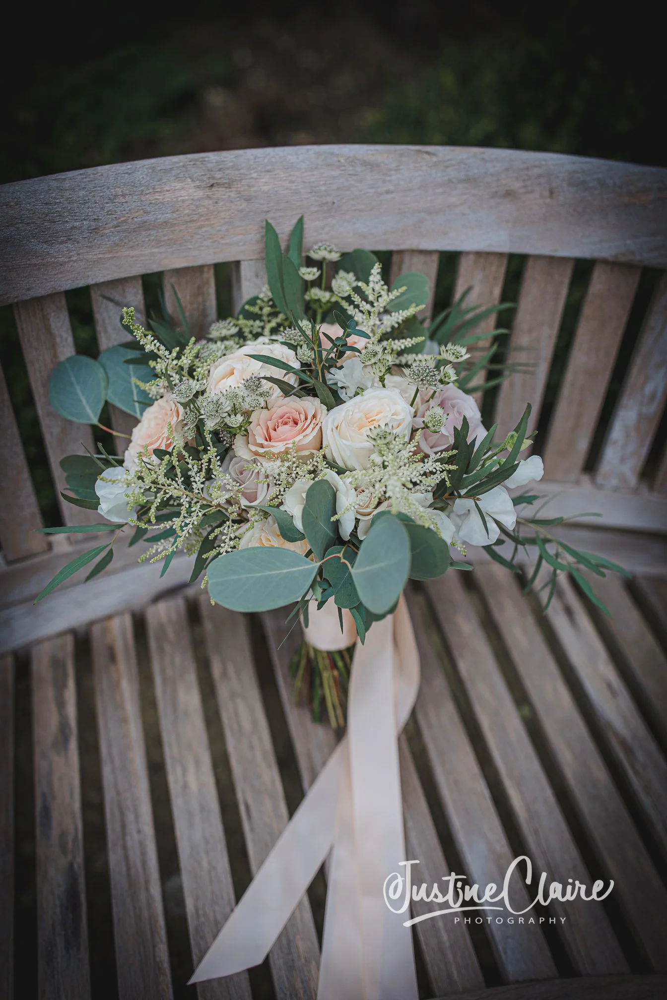 A bouquet of light pink and white roses with greenery and white filler flowers on a wooden bench.