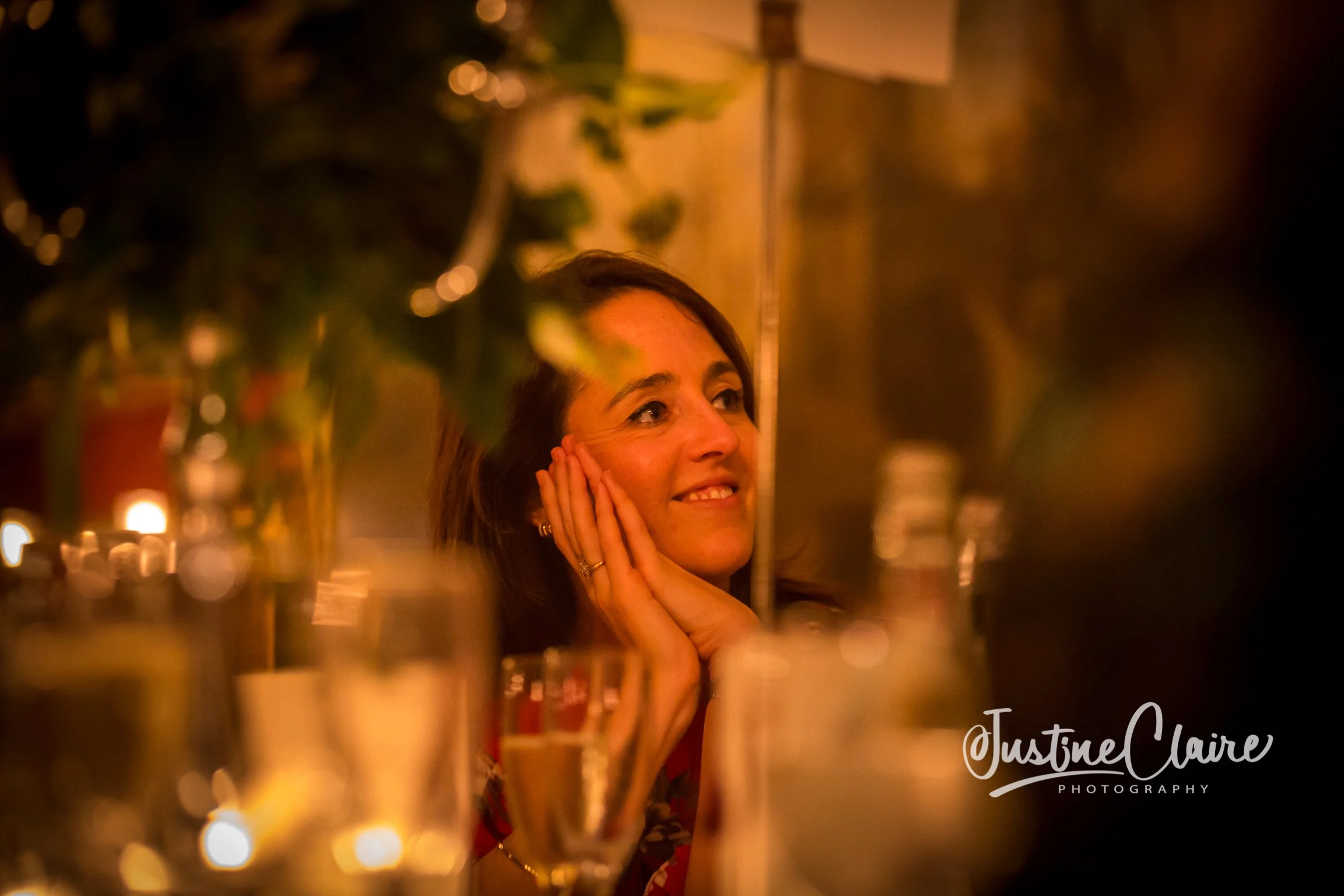 A woman with medium-length dark hair smiling and resting her face on her hands at an event with dim, warm lighting. She is looking slightly to her right, surrounded by blurred glasses, bottles, and decorations.