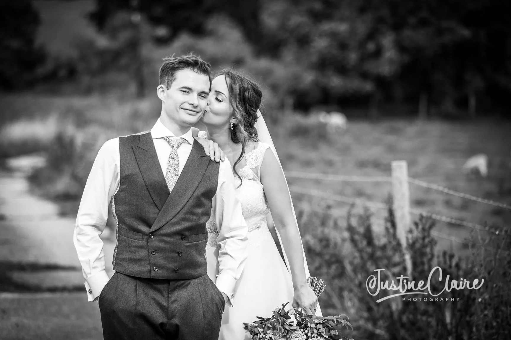 A black and white photograph of a bride kissing a groom on the cheek outdoors, with a rural landscape in the background. The bride is holding a bouquet of flowers and the groom has his hands in his pockets. The photographer's logo, Justine Claire Pho