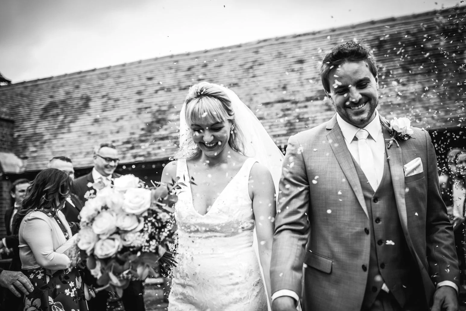 Black and white photo of a newlywed couple smiling as wedding guests throw confetti during their outdoor wedding celebration.