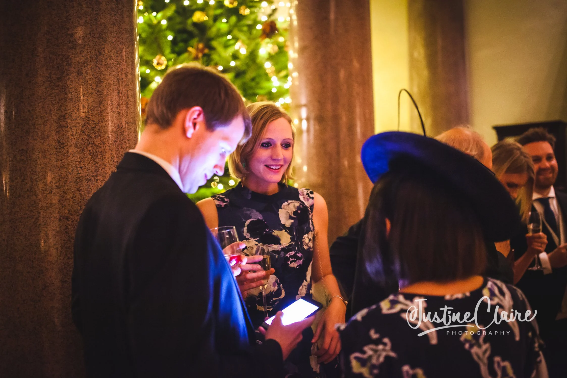 People at a holiday party with a decorated Christmas tree in the background, engaged in conversation, holding drinks, one person looking at a phone, in an elegant venue.