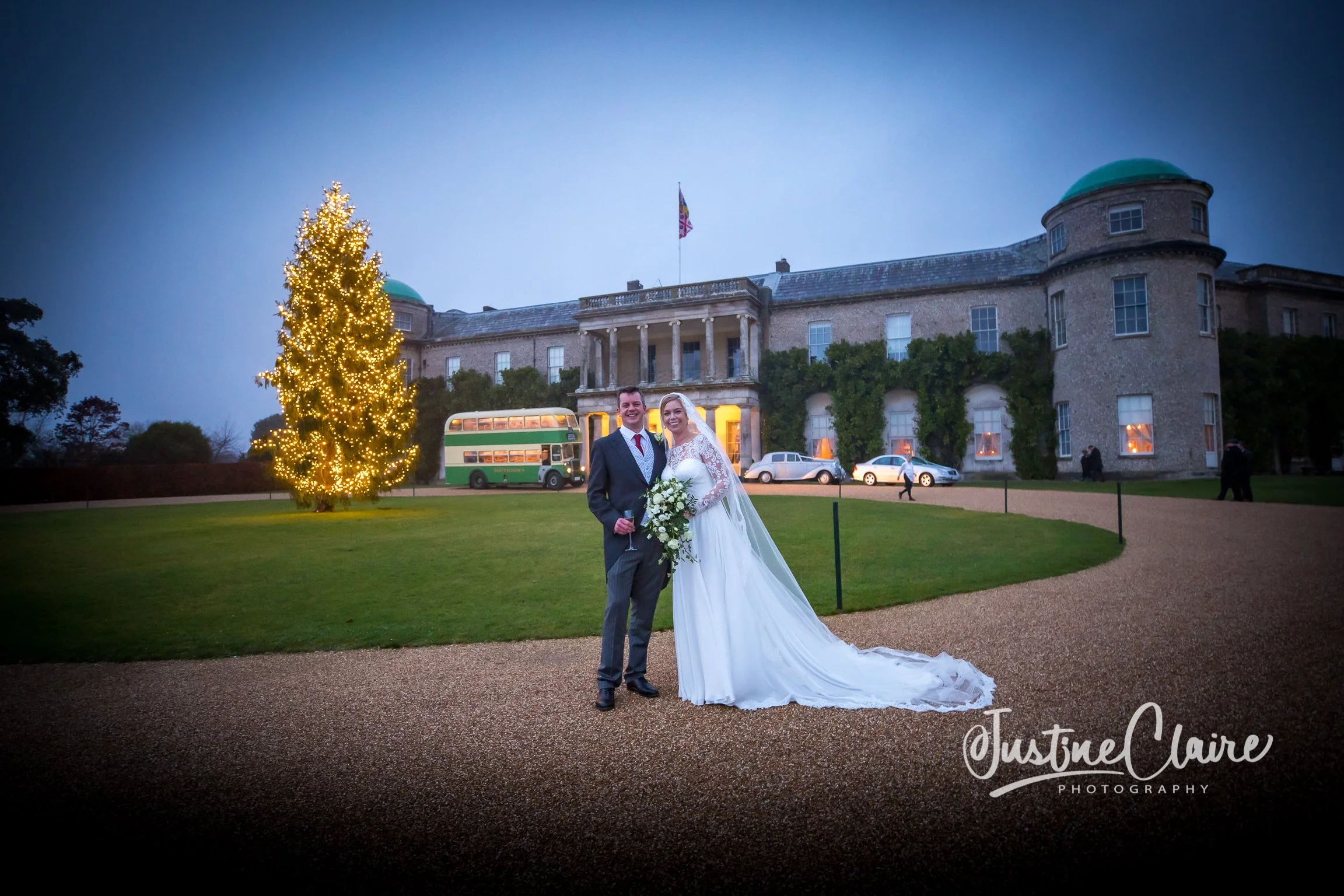 Bride and groom standing outside a large historic building at dusk, wedding celebration, bride in white gown holding bouquet, groom in dark suit, Christmas tree with lights nearby, vintage cars parked, illuminated windows, photographer's watermark.