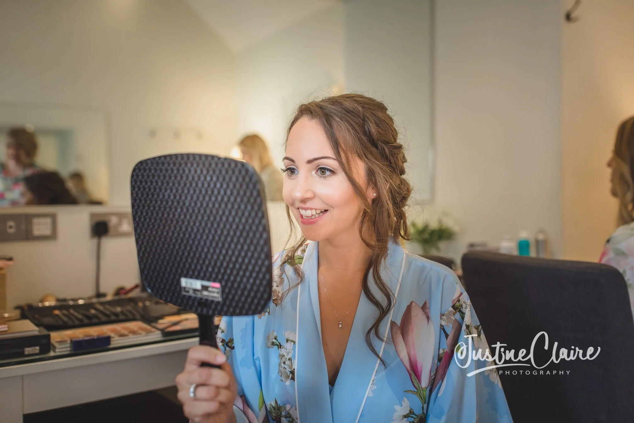 A woman in a floral blue robe is smiling and looking into a handheld mirror. farbridge wedding photography