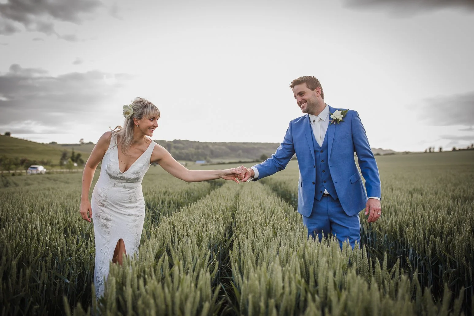 A bride and groom smiling and holding hands in a wheat field during a wedding photoshoot, with cloudy sky and rolling hills in the background.