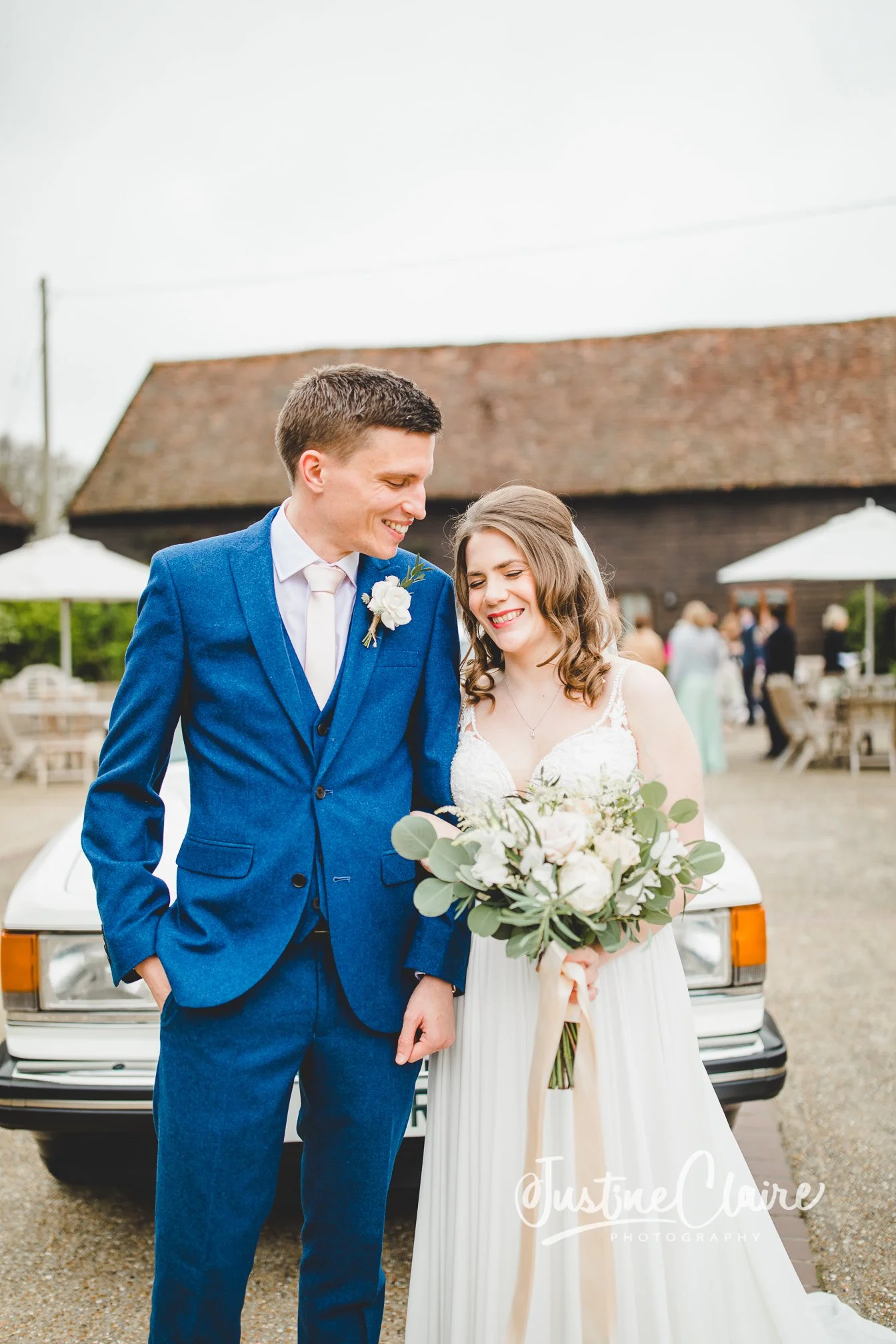 A bride and groom smiling and looking at each other outdoors, standing in front of a white car, with a rustic barn and guests in the background.