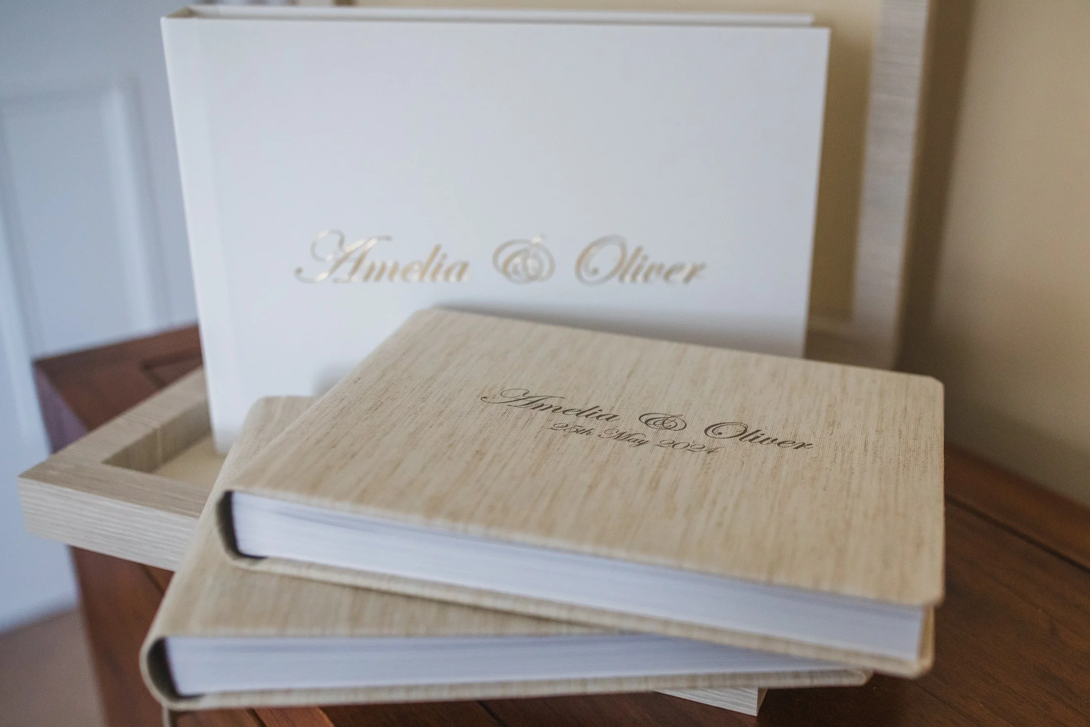 A wedding album with a beige textured cover and inscription, placed on a wooden table in front of a white box with gold script reading from Graphistudio