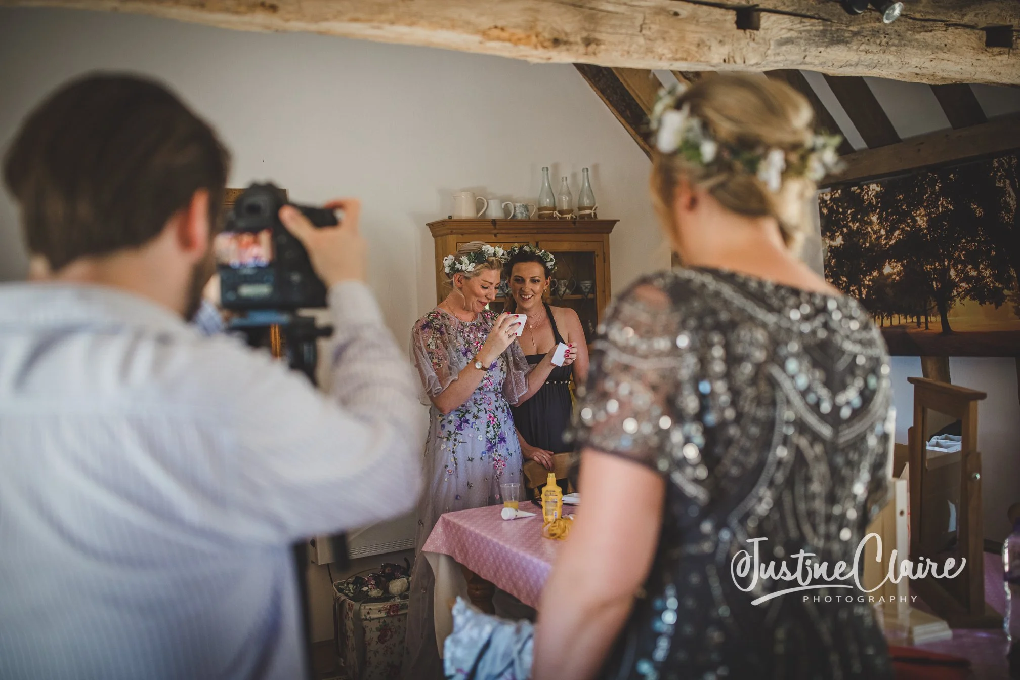 A woman in a floral dress with a flower headband looks at her phone, standing at a table with a pink tablecloth, while two other women, also wearing flower headbands, smile and look on. A man in a white shirt is taking a photo of them with a camera,