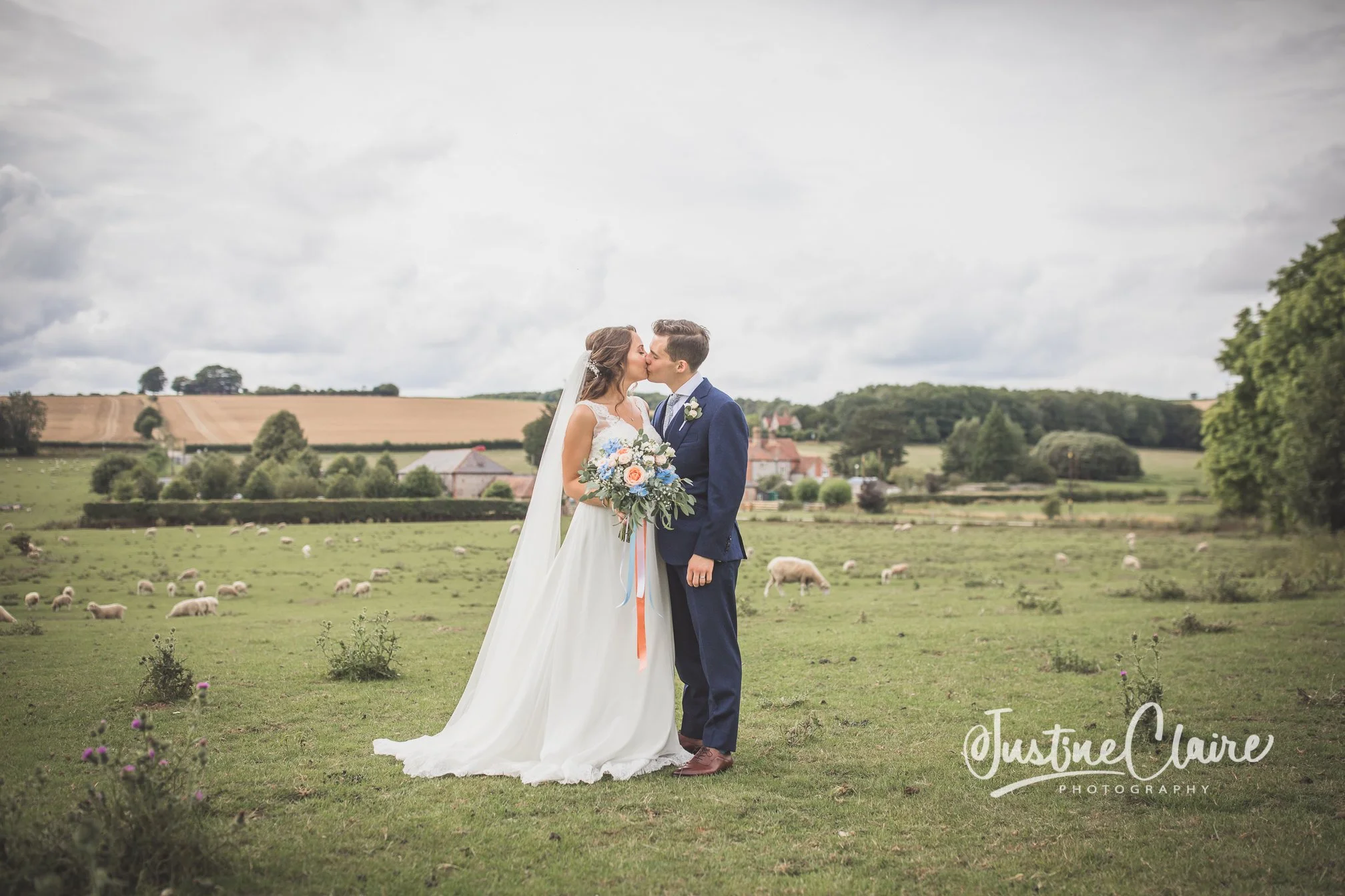 A bride and groom kissing outdoors in a green field with sheep, trees, and houses in the background on a cloudy day. Farbridge gorgeous wedding venue chichester farm buildings