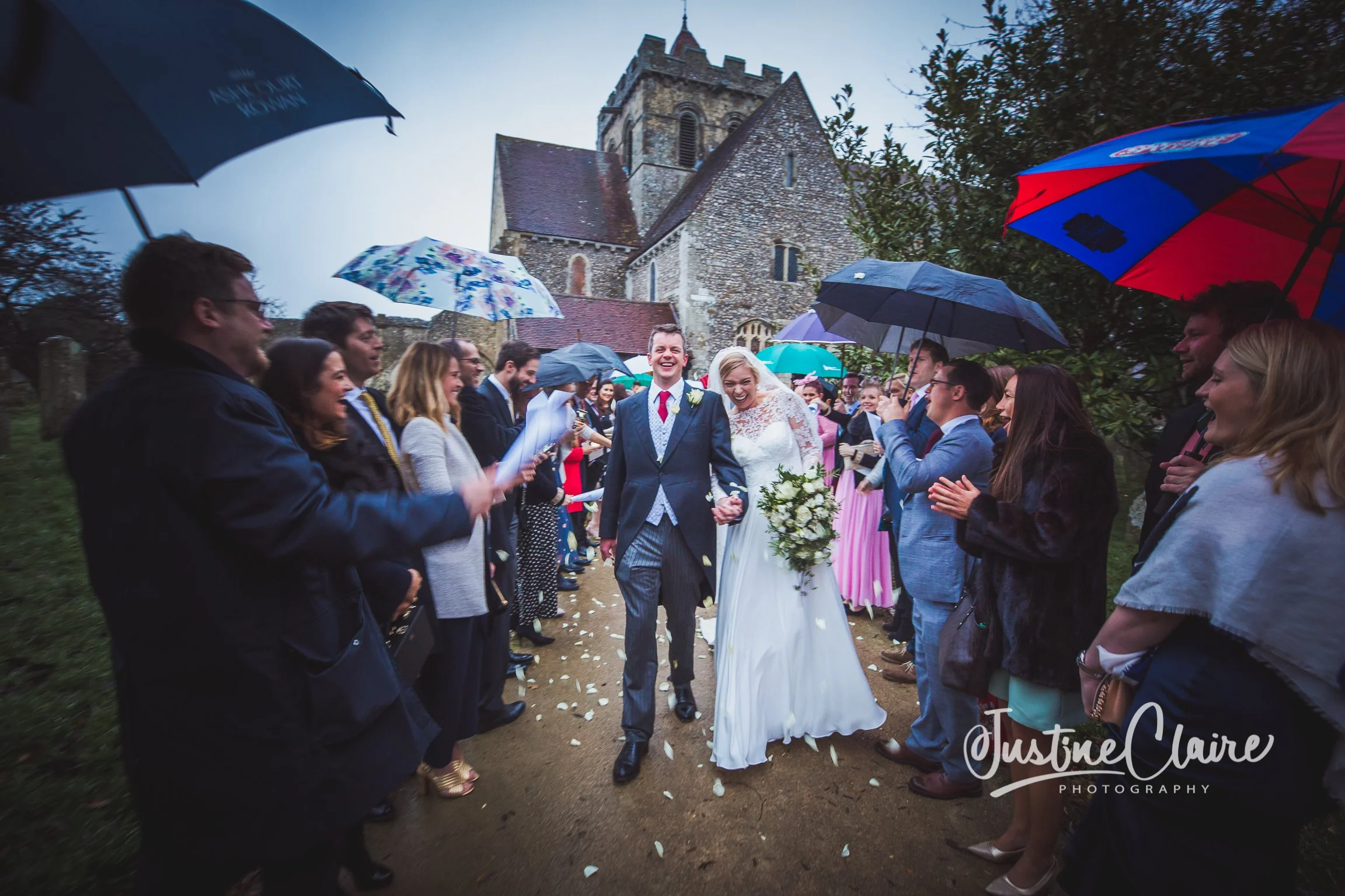 A newlywed couple walking hand in hand and smiling, surrounded by guests holding umbrellas, as they celebrate outdoors near a church during a rainy wedding day.