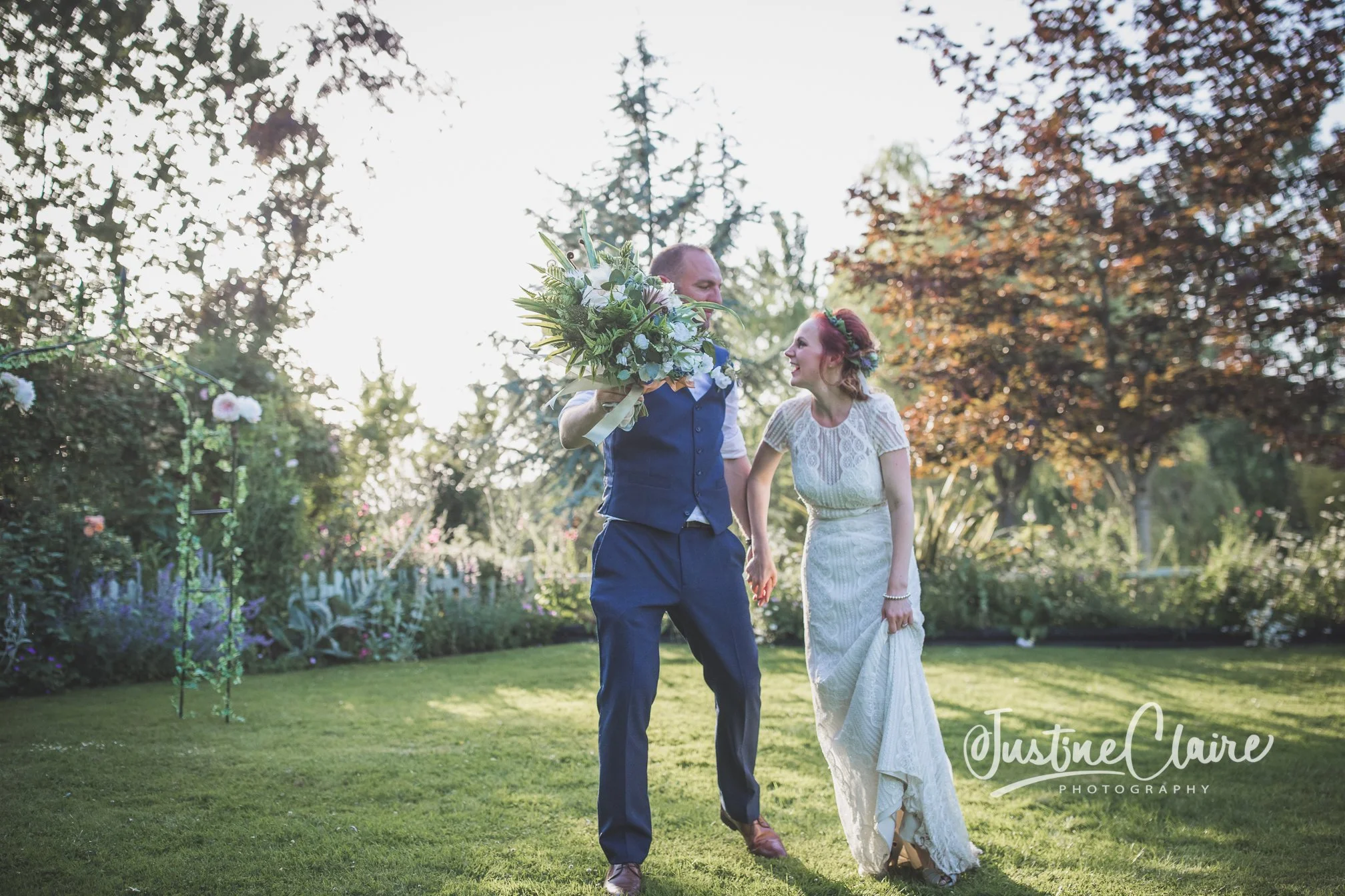 A bride and groom holding hands and walking in a garden during sunset, the groom holding a large bouquet of flowers, with trees and flowering plants in the background.