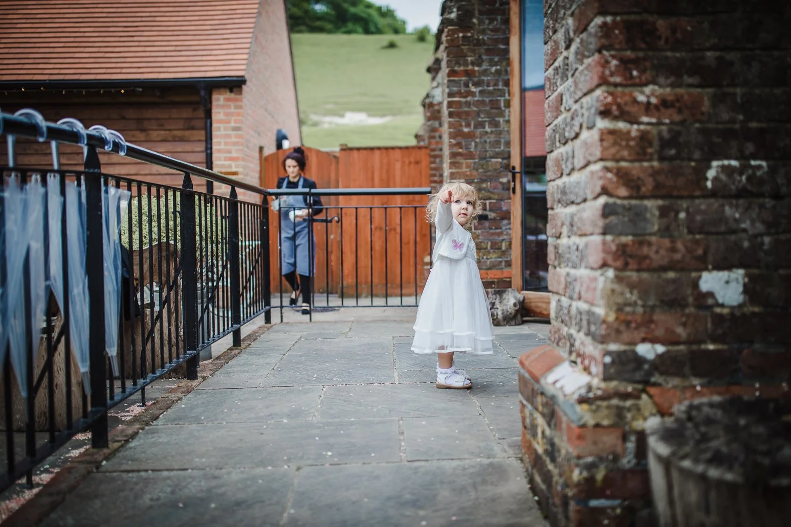 Young girl in a white dress and platform shoes pointing her finger while standing on a paved backyard patio. In the background, a woman with an umbrella and apron is walking past a wooden gate near brick and wooden houses, with a green hillside in th