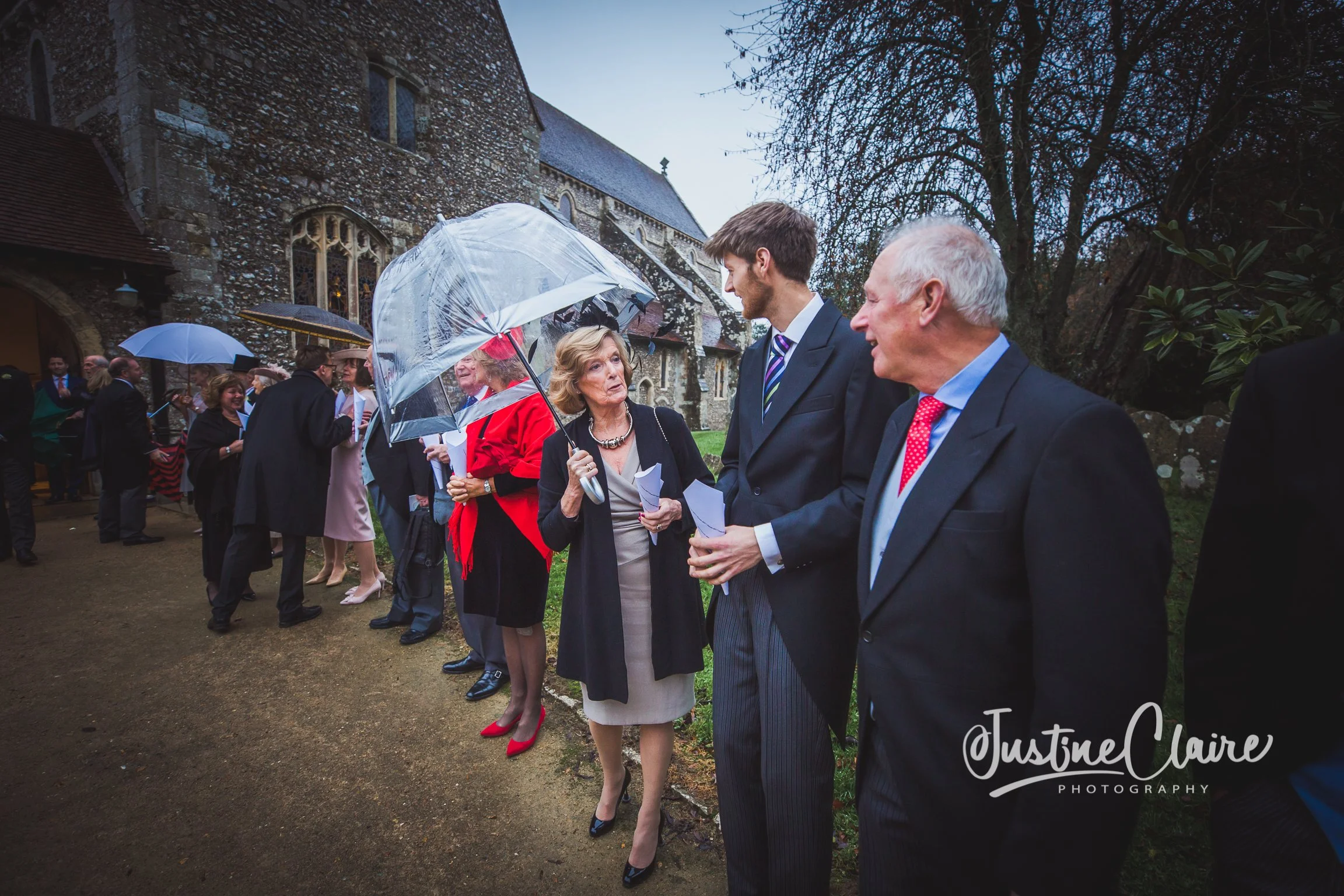 People dressed formally, standing in line outside a church, holding umbrellas on a rainy day.