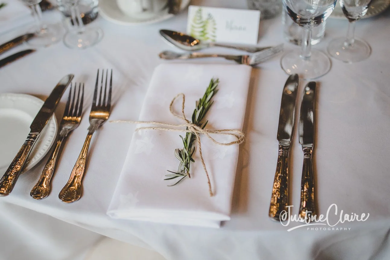Elegant table setting with a white cloth napkin tied with string and a sprig of rosemary, surrounded by silverware and wine glasses.