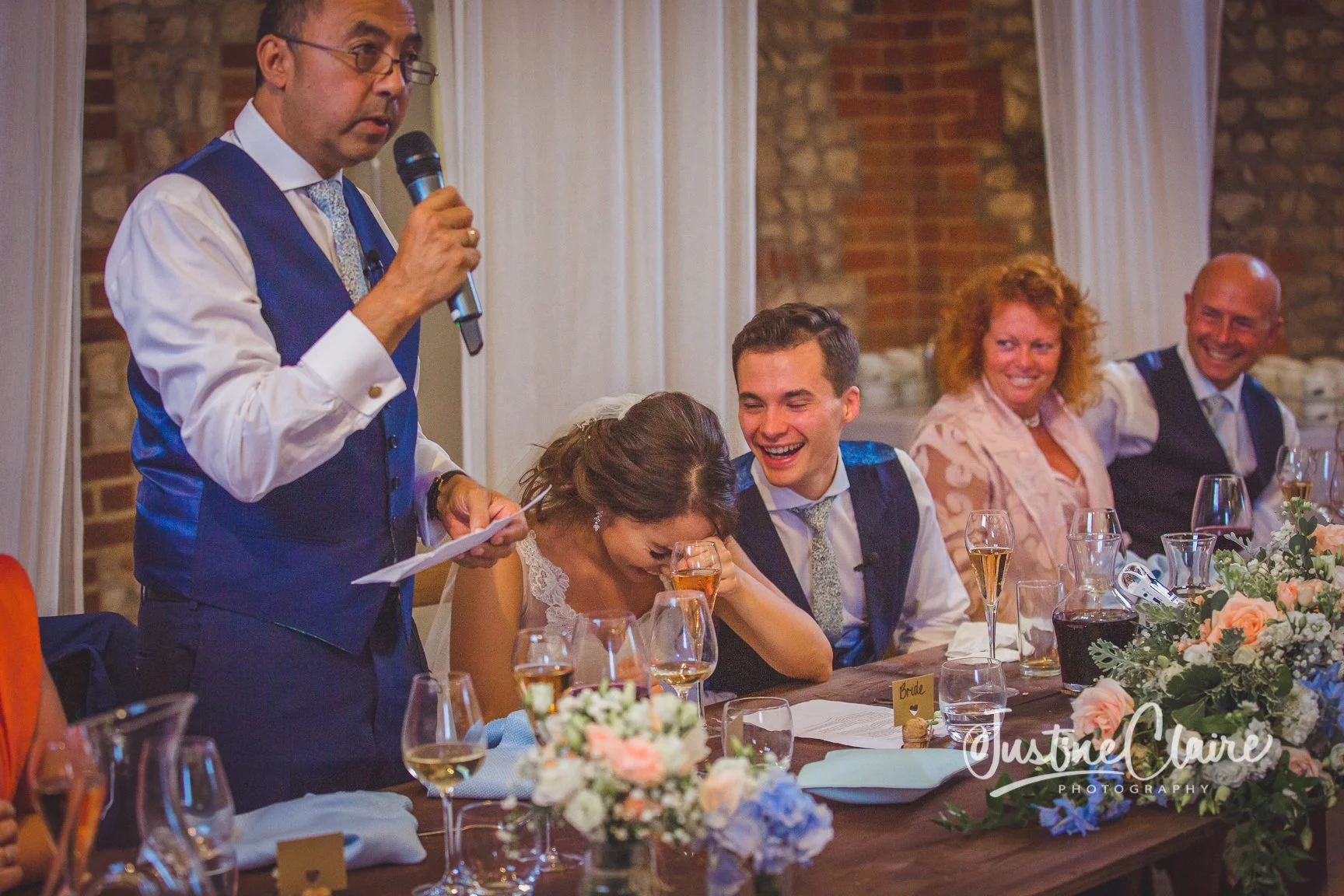A man giving a speech at a wedding reception with a woman laughing and covering her face, seated at a decorated table with wine glasses and a floral centerpiece, surrounded by smiling guests.