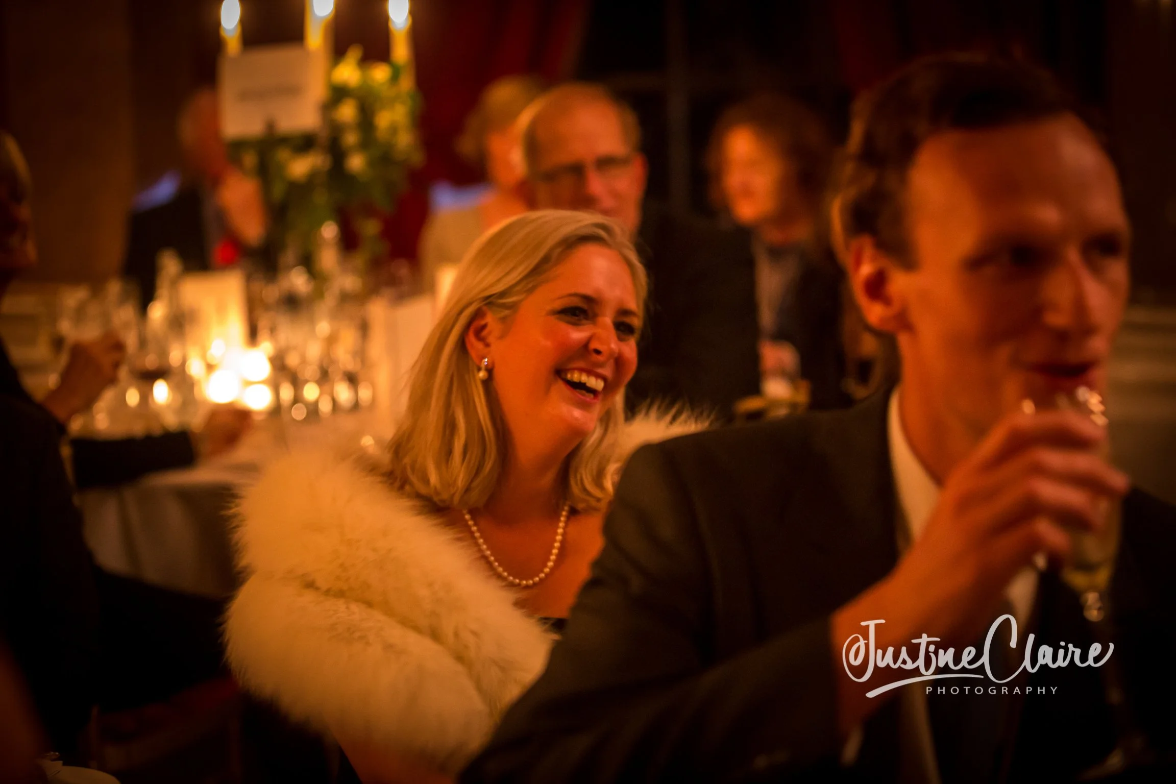 A woman with blonde hair wearing pearl earrings and a fur coat laughing at a formal event, with others in elegant attire in the background