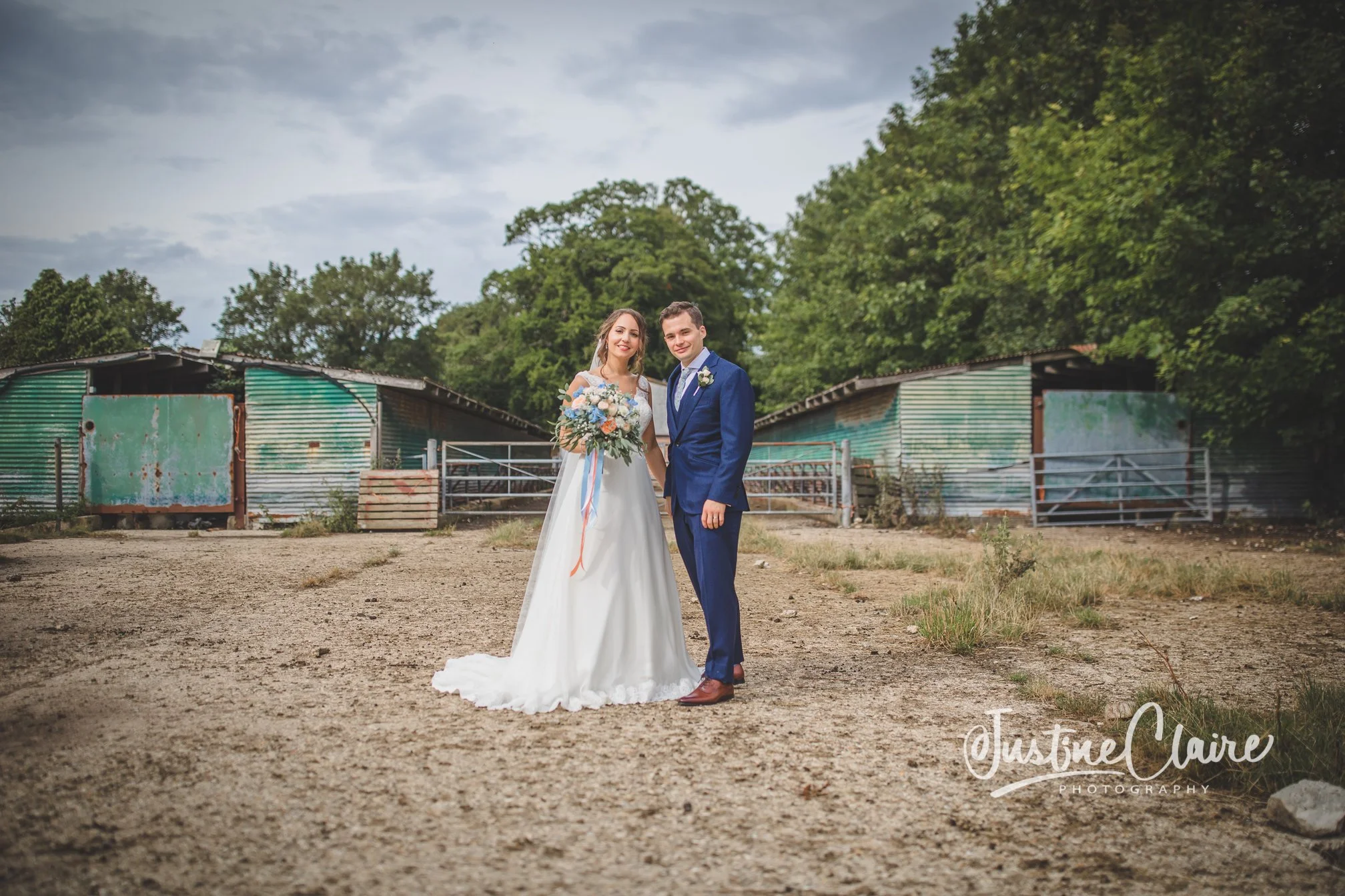 A bride and groom standing on a dirt path in front of a rustic, weathered barn with greenish-blue metal siding. The bride is in a white wedding dress holding a bouquet, and the groom is in a blue suit with a boutonniere. Green trees and a cloudy sky