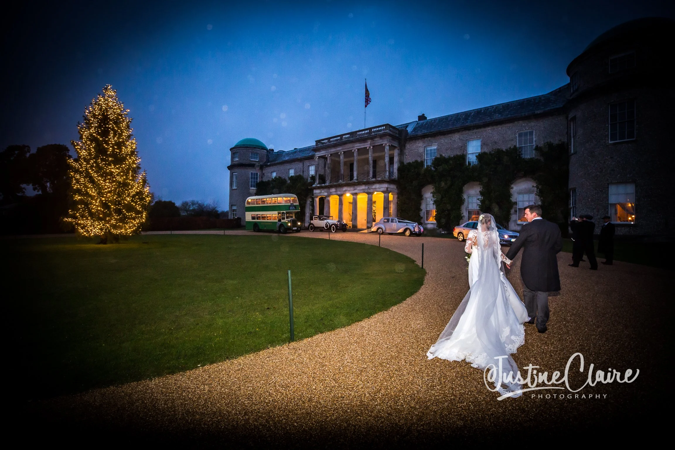 Wedding couple walking on a gravel path outside a large vintage mansion at night, with parked cars, a lit Christmas tree, and a double-decker bus in the background.