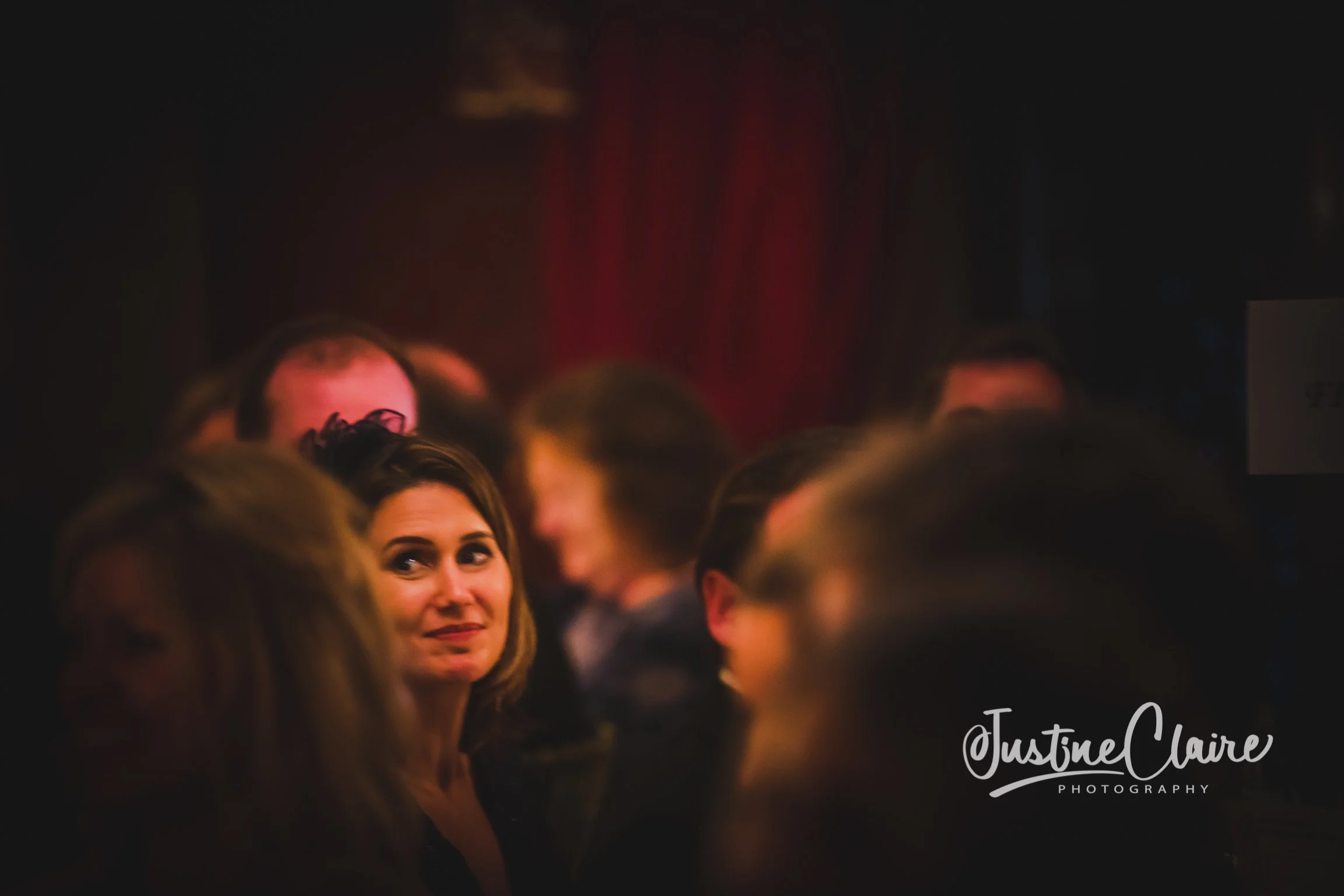 A woman with shoulder-length brown hair and a slight smile, looking to the side in a dimly lit setting with other people around her, possibly at a formal event or gathering.