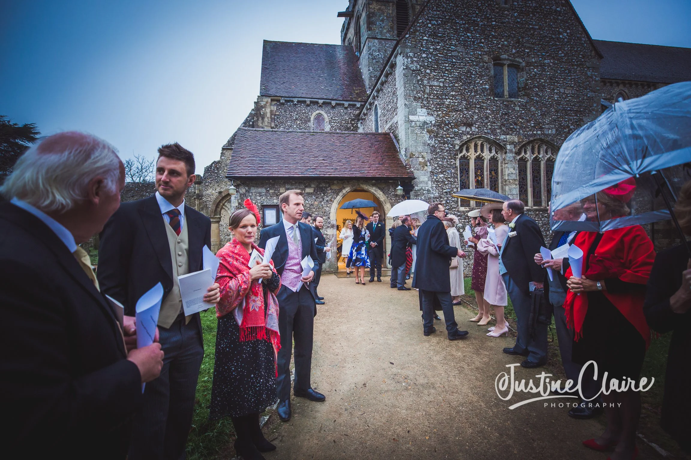 People gathered outside a stone church holding umbrellas on a rainy day, waiting for a wedding ceremony to begin.