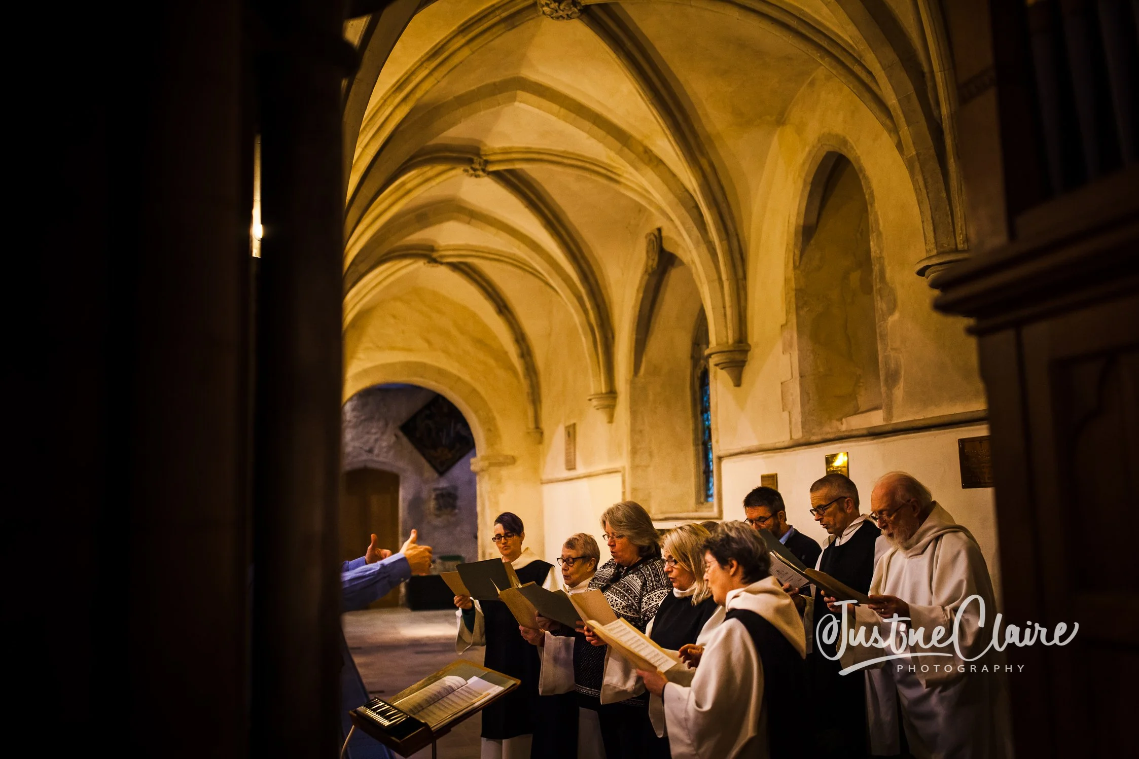 A choir group singing inside a church with stone arches. The conductor's hands are visible, leading the choir members who hold sheet music.