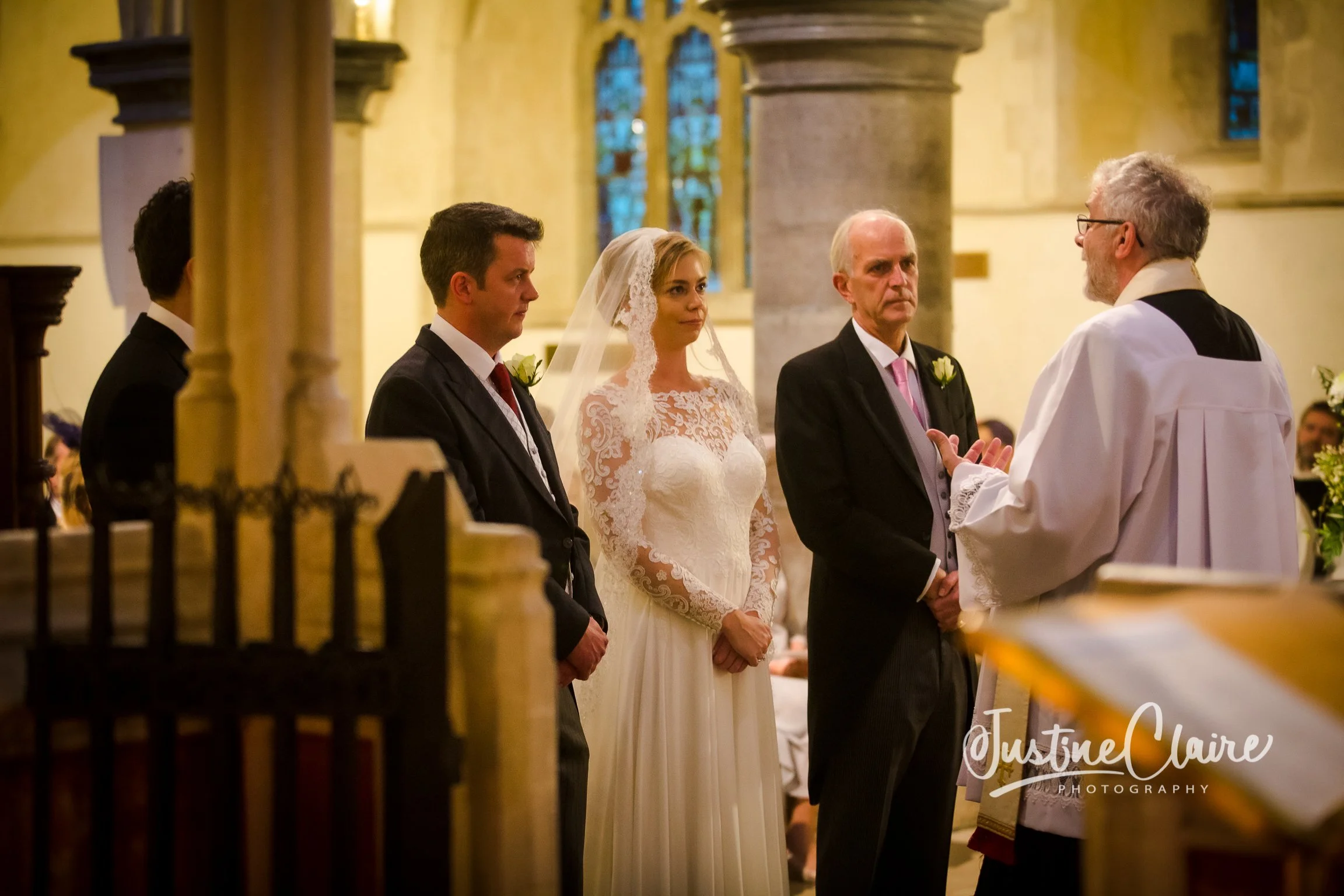 A bride and groom stand before a priest during a wedding ceremony inside a church, with two other men in suits. The bride is wearing a white lace wedding dress with a veil, and the groom is wearing a dark suit. The priest is dressed in white liturgic