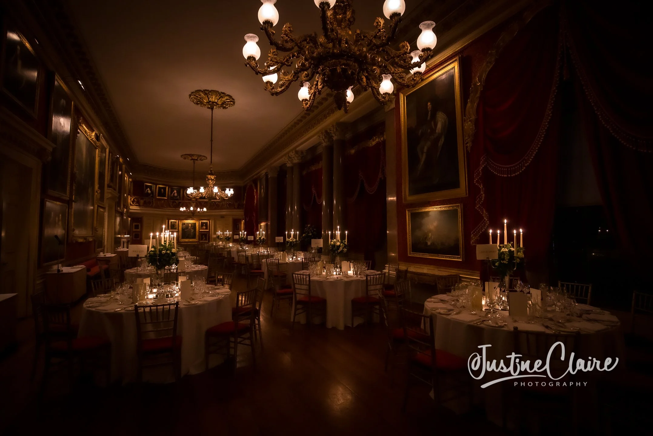 Elegant dining room with round tables covered in white tablecloths, set with glassware and plates, surrounded by wooden chairs with red cushions, chandeliers and candlelit fixtures, gold-framed paintings on red velvet drapes, and dim ambient lighting