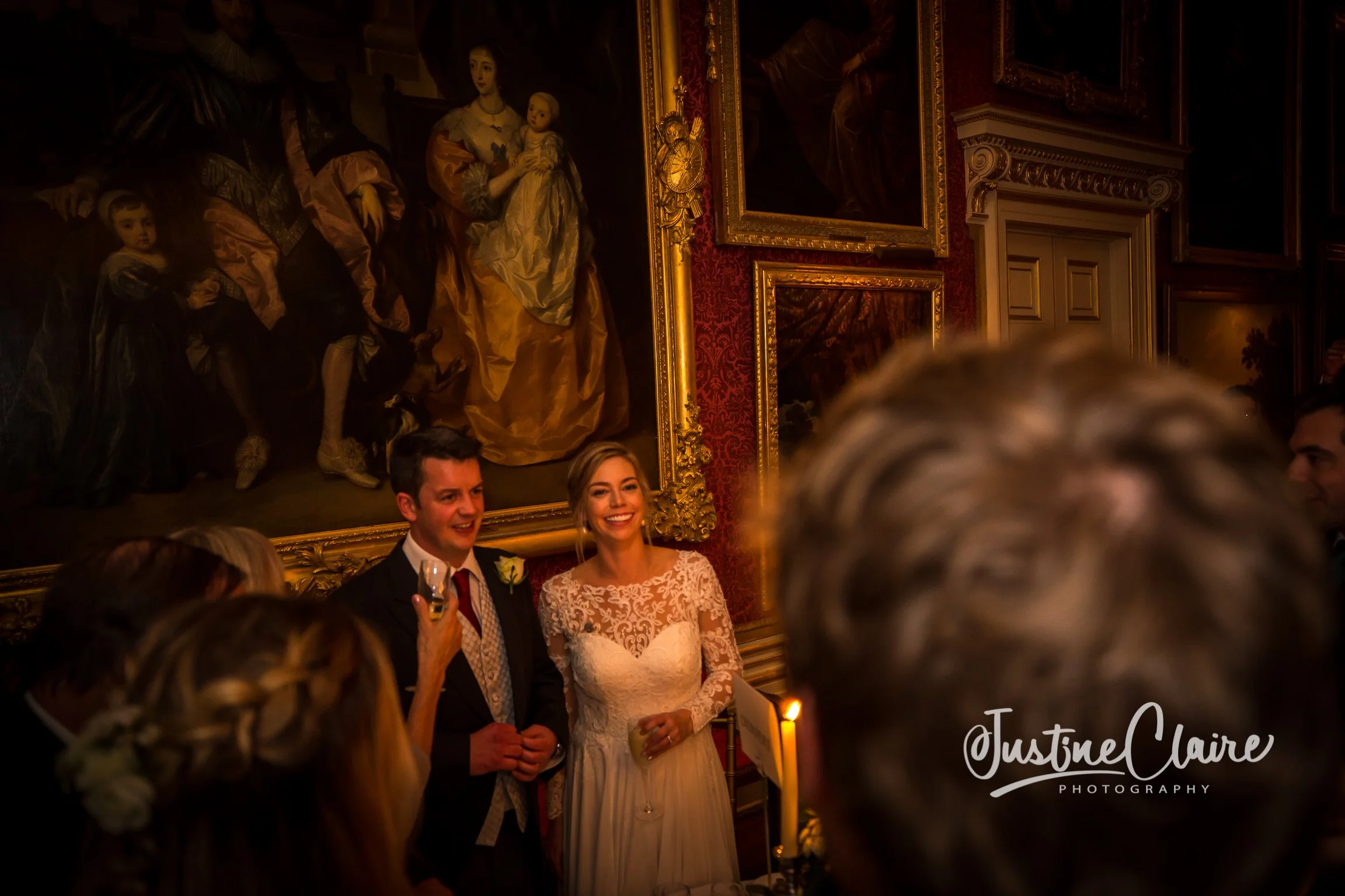 A wedding celebration with a bride and groom smiling, surrounded by guests, in an elegant room with large classical paintings and ornate gold frames.