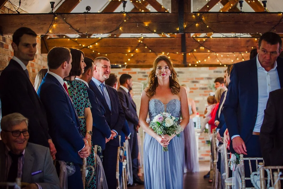A bride in a silver lace dress holding a bouquet of white and purple flowers, walking down the aisle during a wedding ceremony inside a rustic venue with string lights and wooden beams.