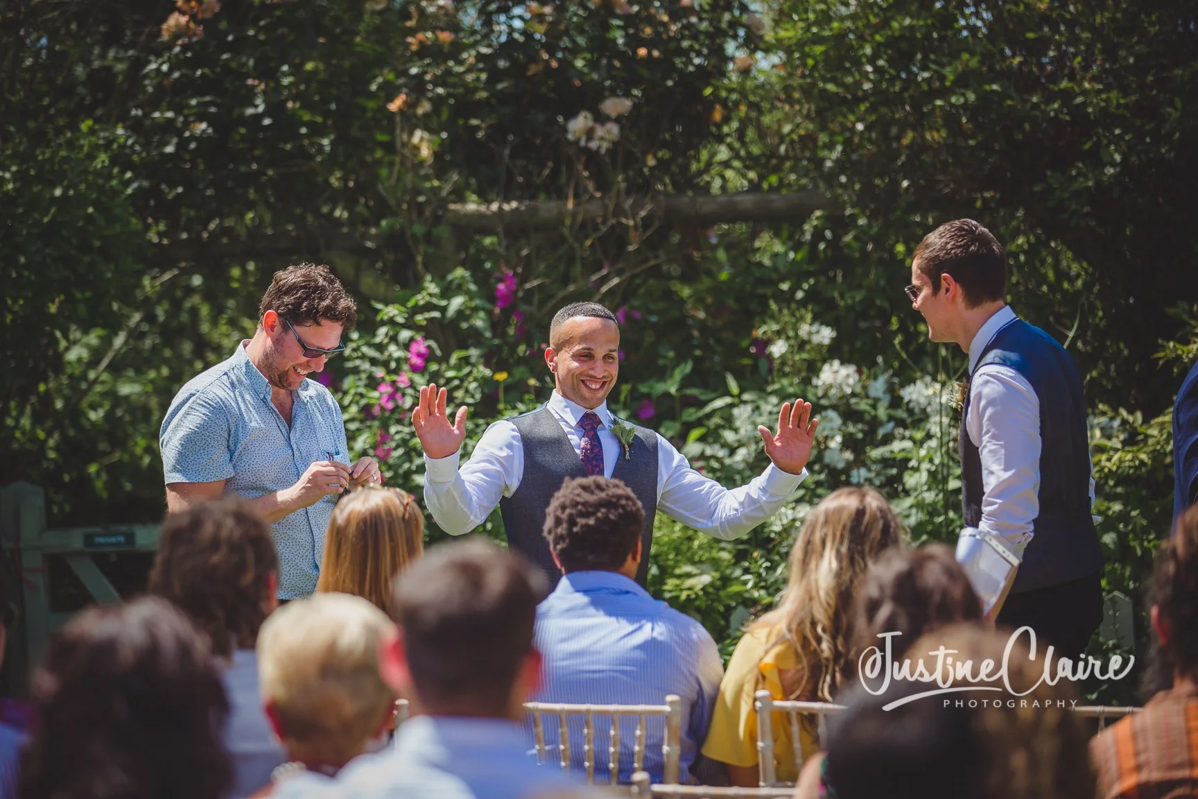 A gay wedding ceremony outdoors with two men standing at the altar, surrounded by guests, with a man in a blue shirt and another man in a navy vest and white shirt scheduling the vows. The groom in a grey vest and floral tie is smiling with his hands