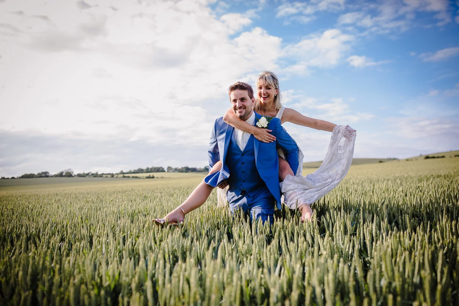 A bride and groom in wedding attire enjoying time together in a field of wheat, with a partly cloudy sky in the background.