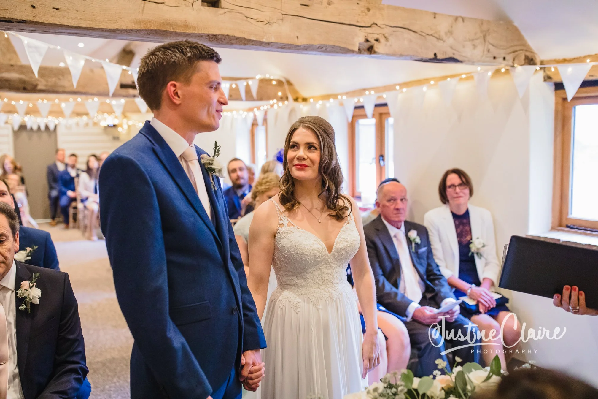 Bride and groom holding hands during their wedding ceremony surrounded by seated guests in a decorated indoor venue. Fitzleroi barn