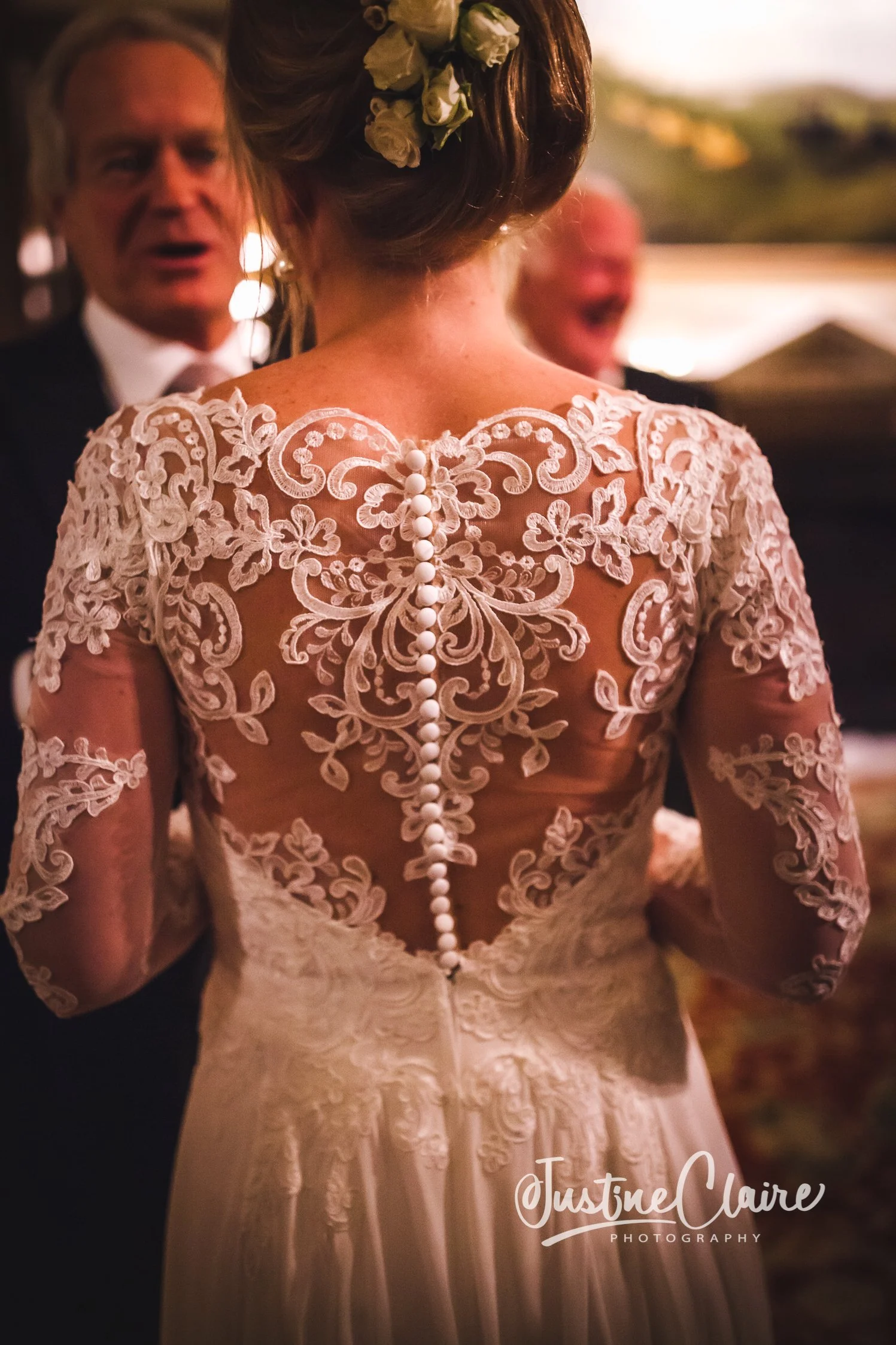 Back view of a bride in a lace wedding dress with a row of fabric-covered buttons, attending a wedding reception outdoors during sunset. Two guests are visible in the background, one man in a suit and another person smiling.
