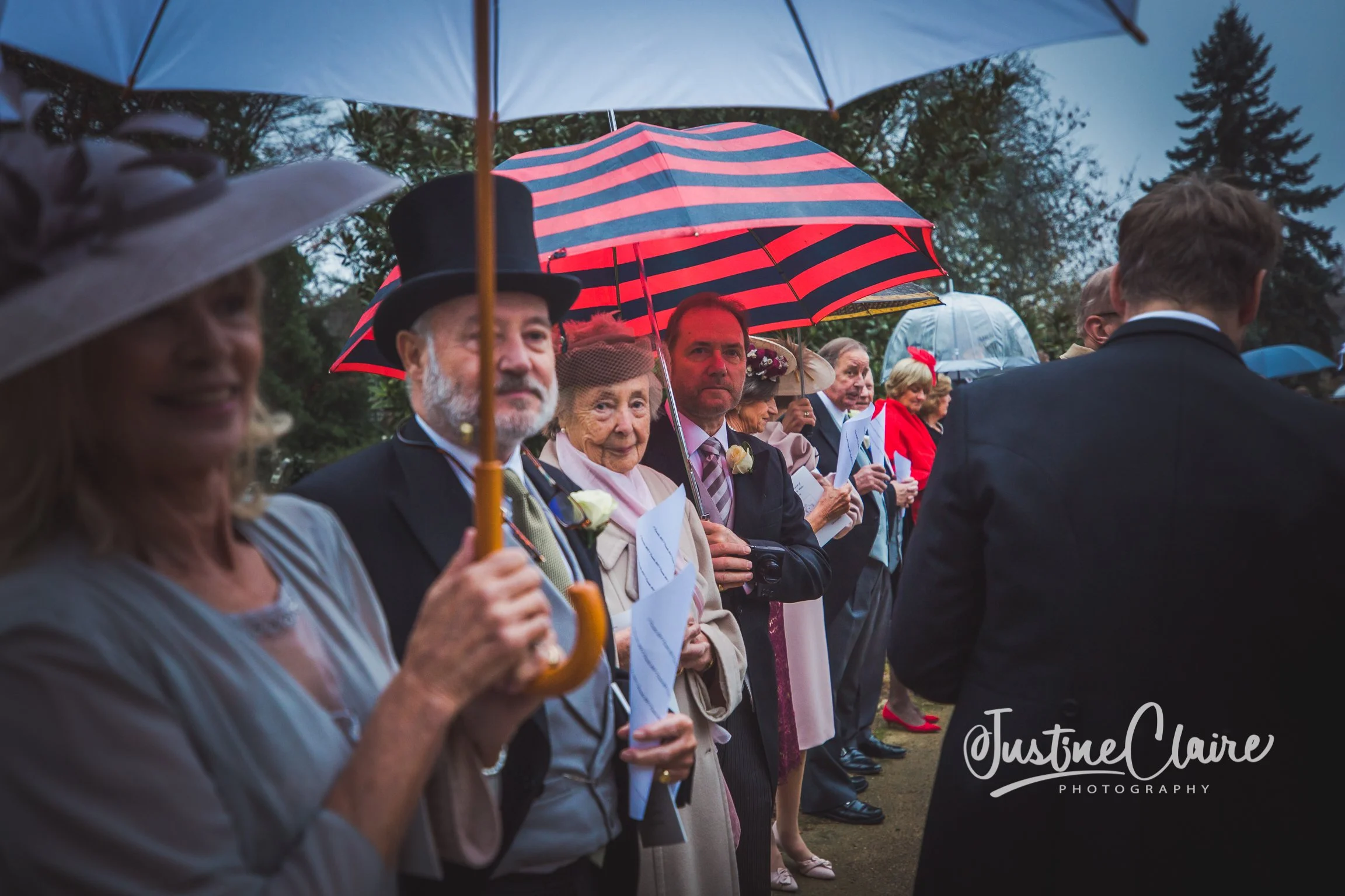 People gathered outdoors at a wedding ceremony holding umbrellas in rainy weather, with some dressed in formal attire and wearing corsages.