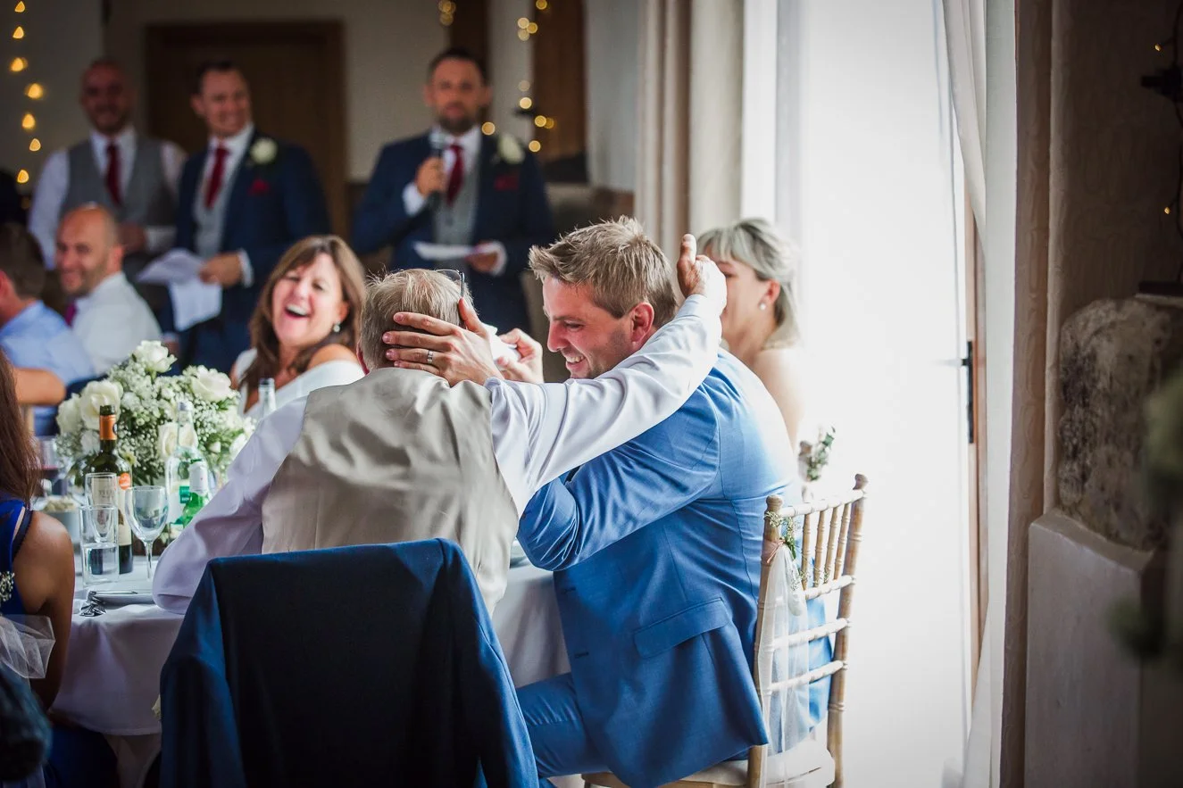 At a wedding reception, a man in a blue suit is playfully hugging an older man, both smiling and sharing a joyful moment, with several guests in the background enjoying the celebration.