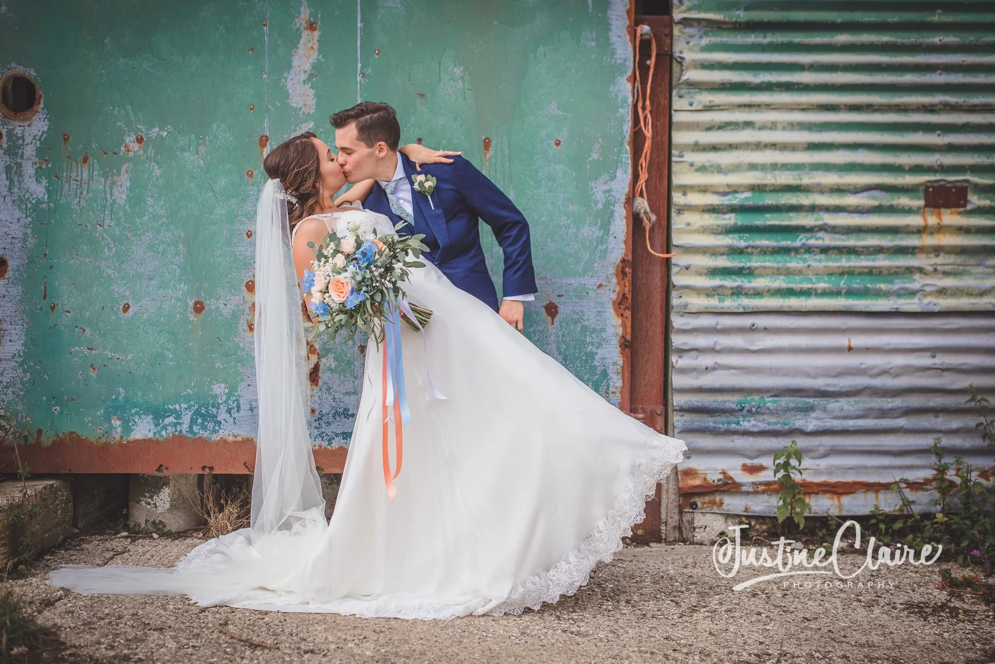 A bride and groom kissing in front of a weathered green and rusted metal wall. The bride is wearing a white wedding dress and holding a bouquet of flowers. The groom is dressed in a navy blue suit.